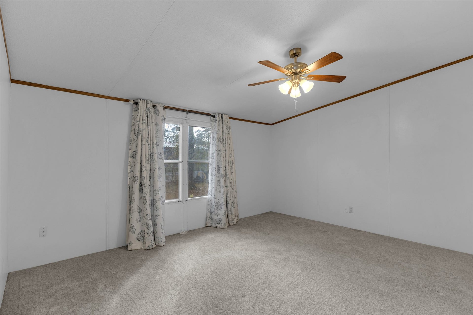 Den room offers a generous carpeted space with white walls and a ceiling fan. Sunlight streams through the large window with patterned curtains, enhancing the airy and welcoming atmosphere.