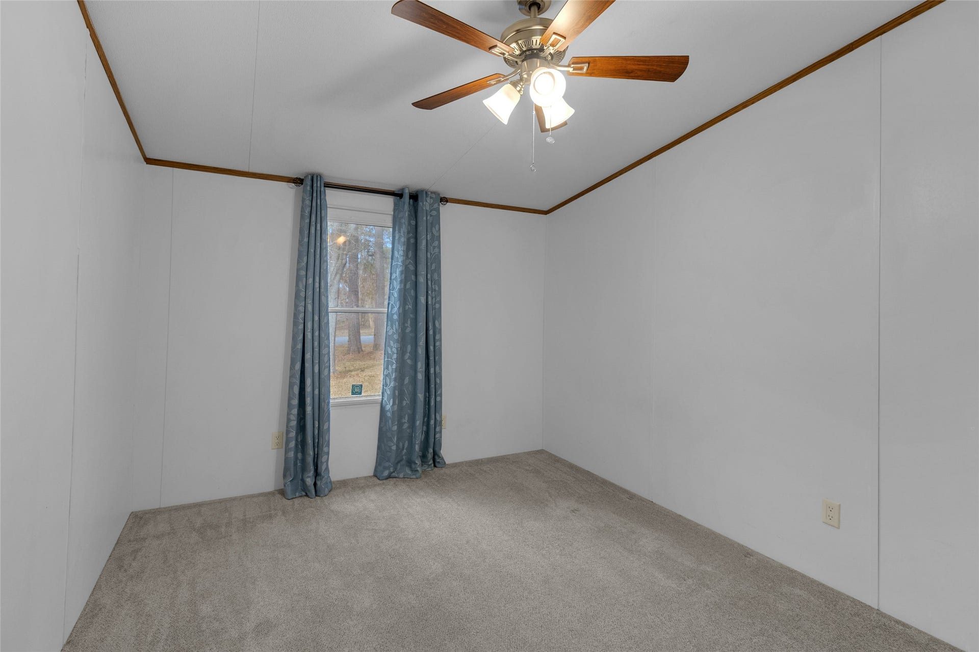 Second Bedroom features neutral-colored walls and carpet, a ceiling fan with lights, and a window with blue curtains, providing a cozy and simple space.