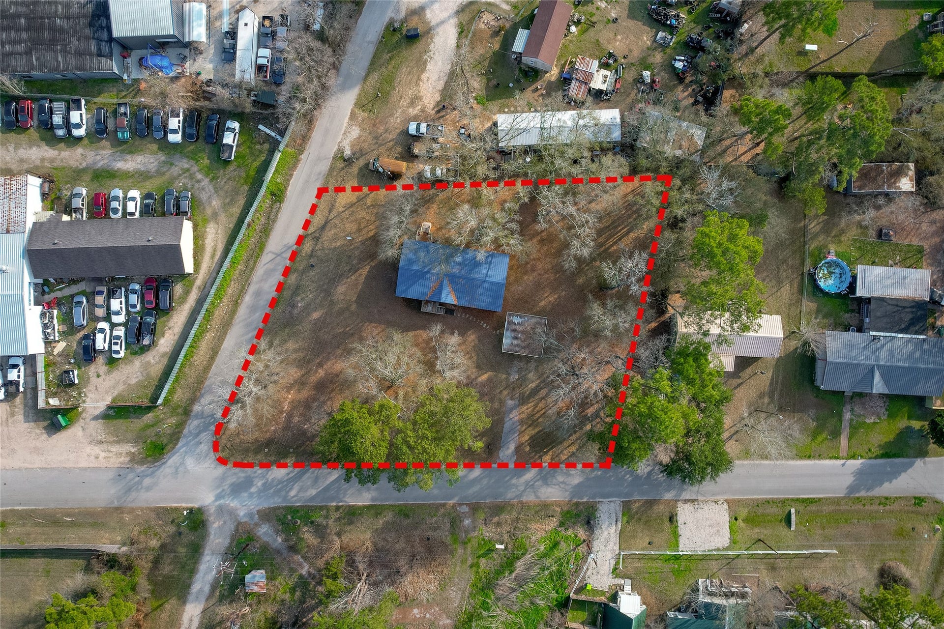 This aerial photo shows a property outlined in red, featuring the house with a blue roof, surrounded by trees. It's located on a corner with access to two roads and is adjacent to a developed area with several buildings and parked cars. The lot is spacious, offering potential for development.