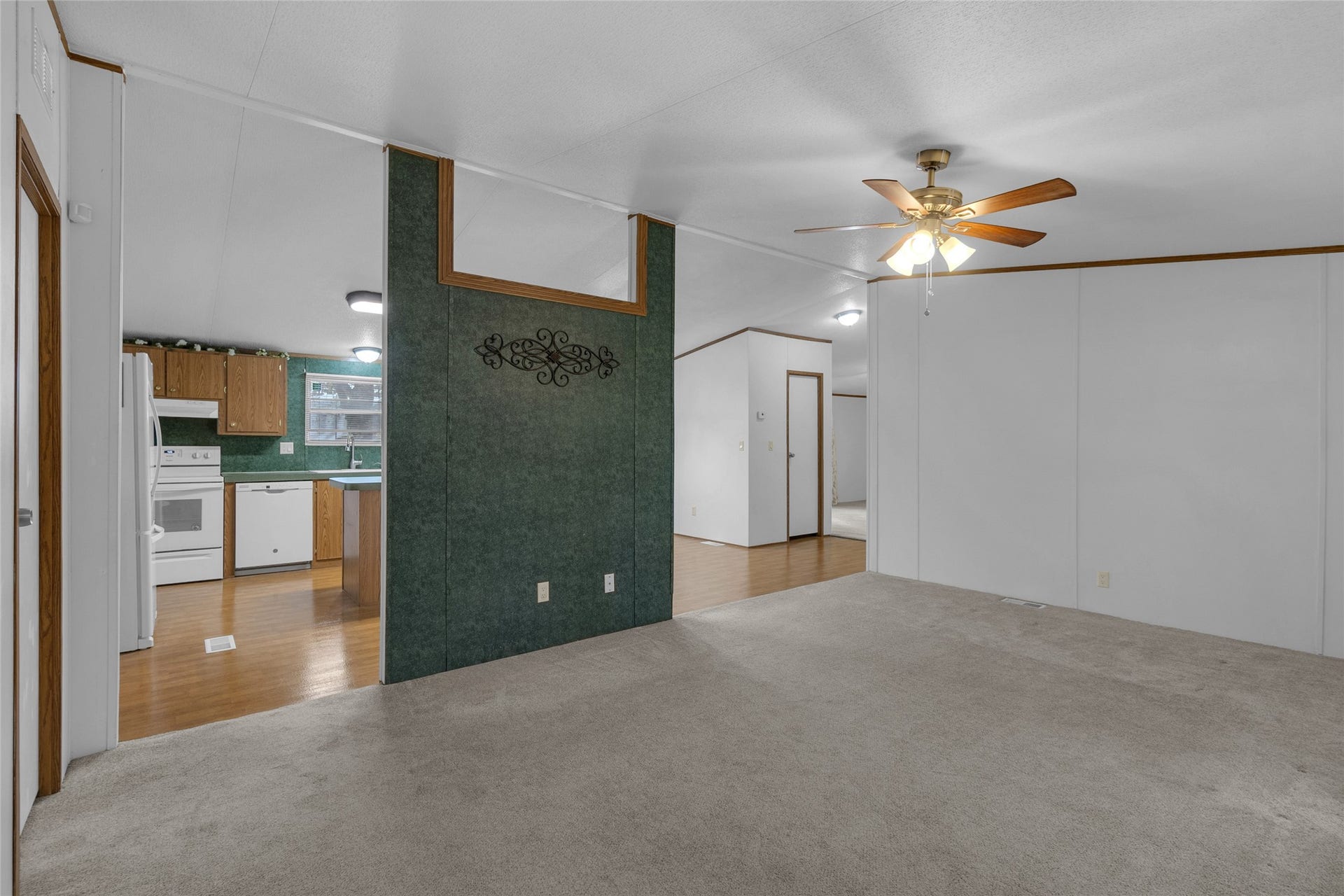 This photo features an open living space with a ceiling fan, neutral carpet, and an adjacent kitchen with wood flooring and appliances. The area has clean, white walls and a decorative green accent wall, creating a warm and inviting atmosphere.