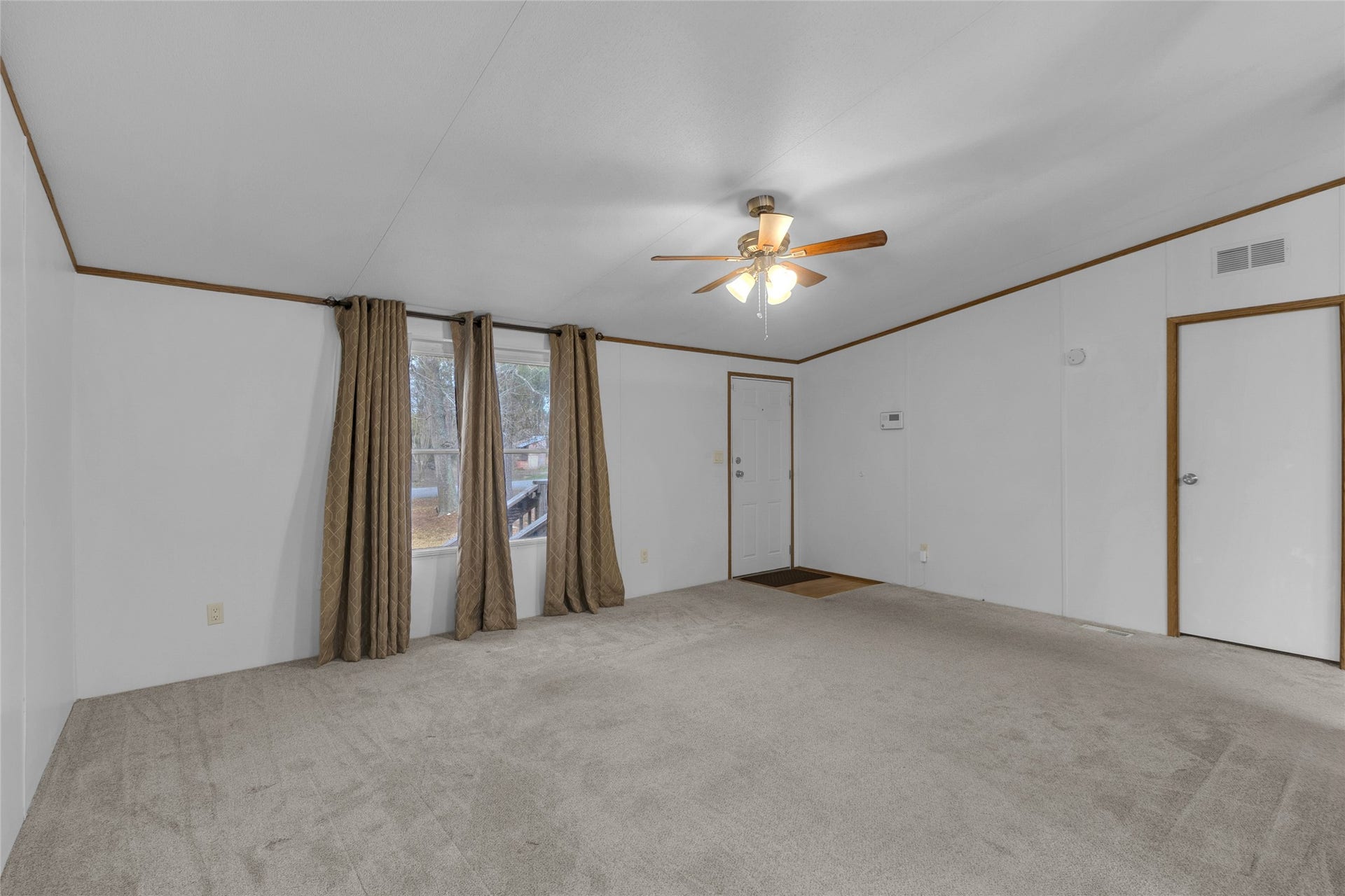 This room features neutral carpeting, white walls with wooden trim, a ceiling fan with lights, and large windows with curtains. It has both an entry door and an interior door, offering good natural light.
