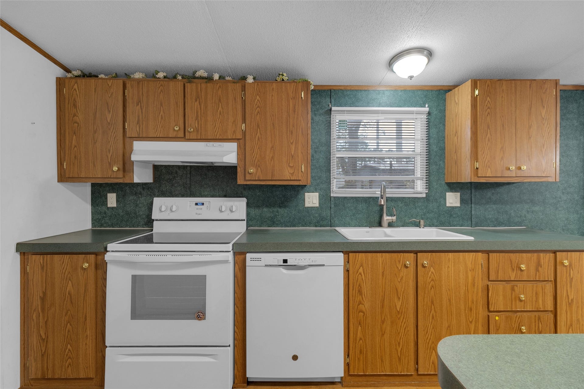 This kitchen features wood cabinets, a white electric stove, and dishwasher, with a green countertop formica. It has a window above the sink for natural light and a ceiling light fixture.