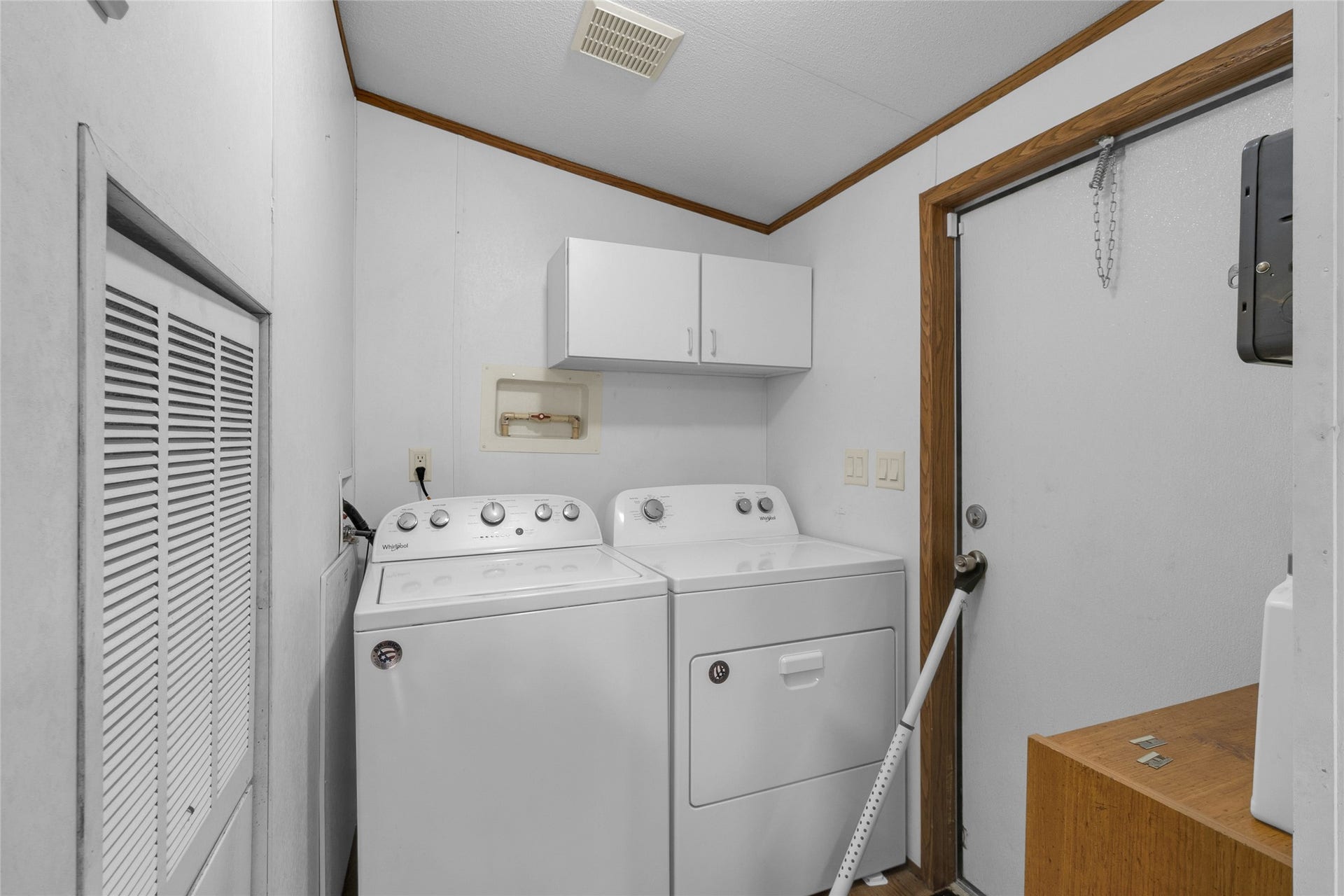 This photo shows a compact laundry room equipped with a washer and dryer. It features overhead storage cabinets and a door leading outside, with white walls and wood trim providing a clean, functional space.