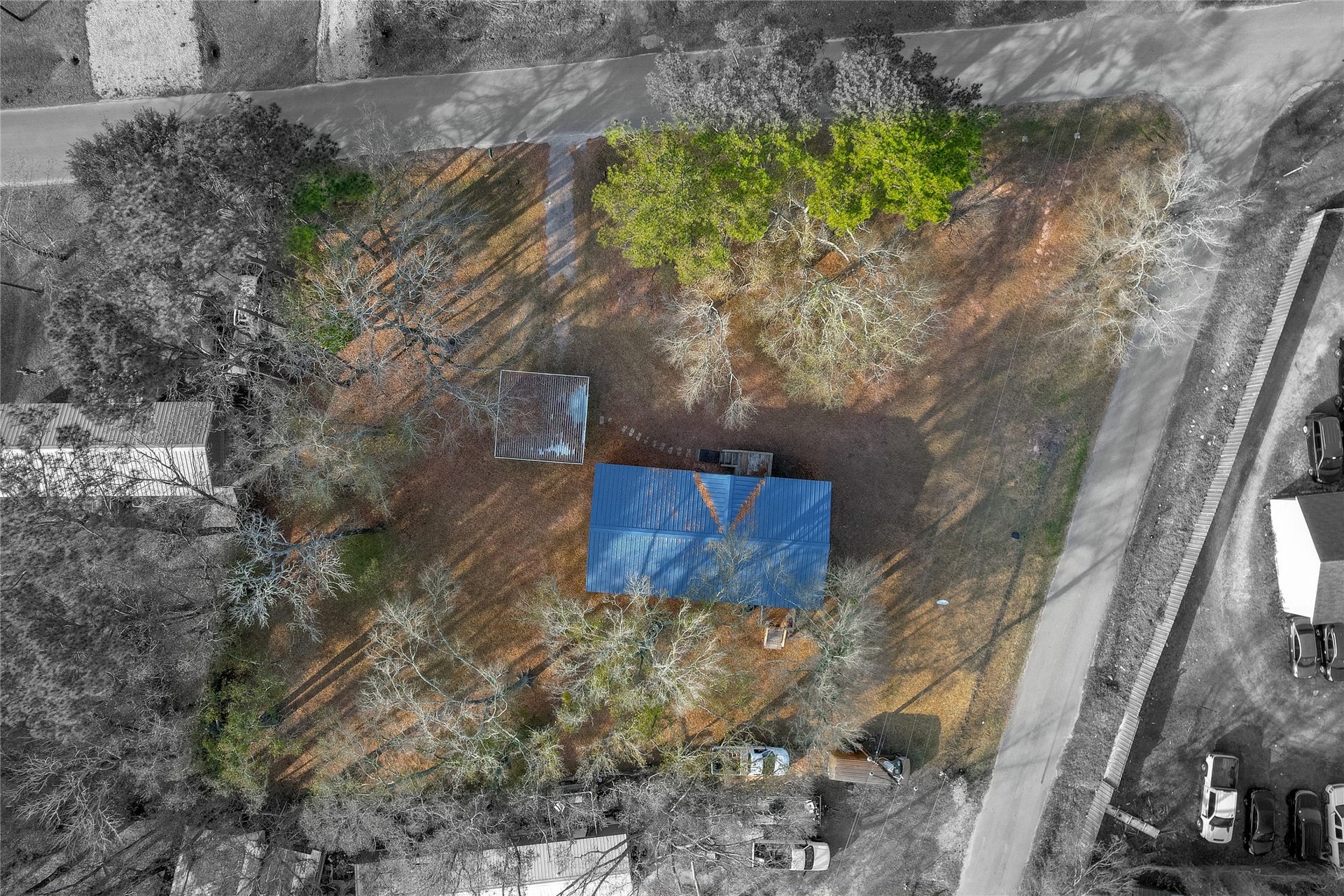 This aerial view shows a property with a blue-roofed house, surrounded by trees and open yard space.