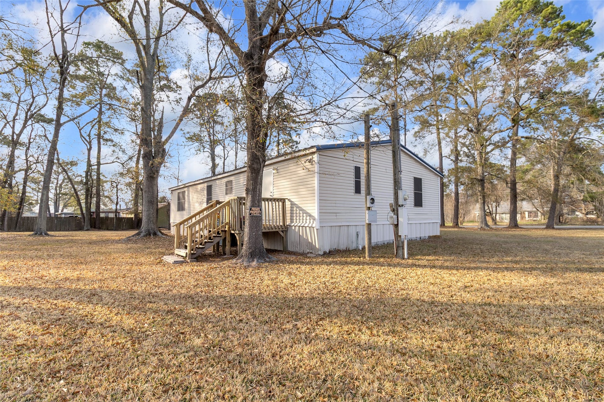 This photo shows a mobile home situated on a spacious, grassy lot with mature trees. The exterior is simple, featuring a small wooden entry backdoor deck. Ideal for those seeking tranquility and outdoor space.