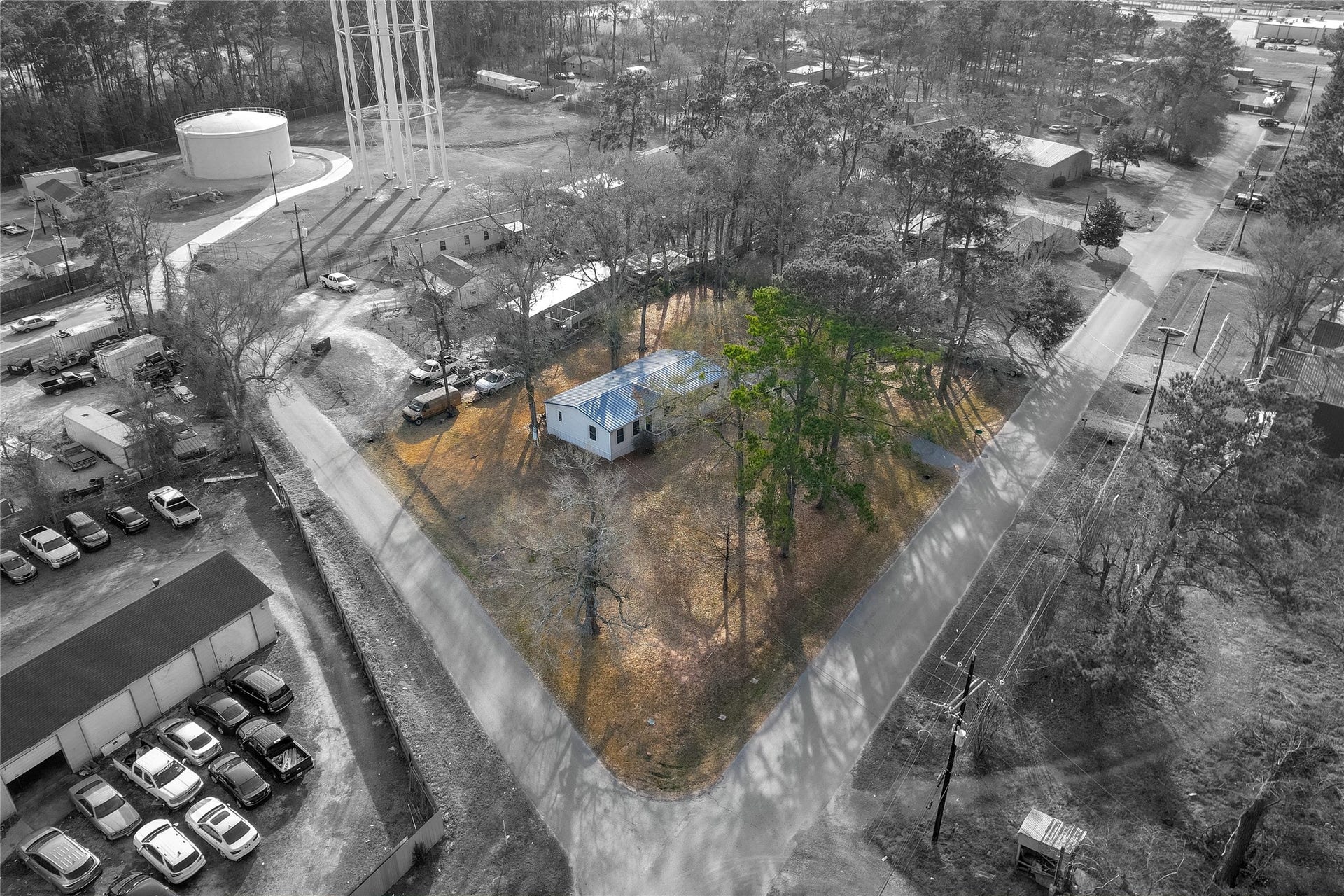 The property is surrounded by tall trees, providing some shade and privacy. Nearby, there are various structures, including a large water tower and some industrial buildings. The area appears to be semi-rural with a mix of residential and commercial properties.