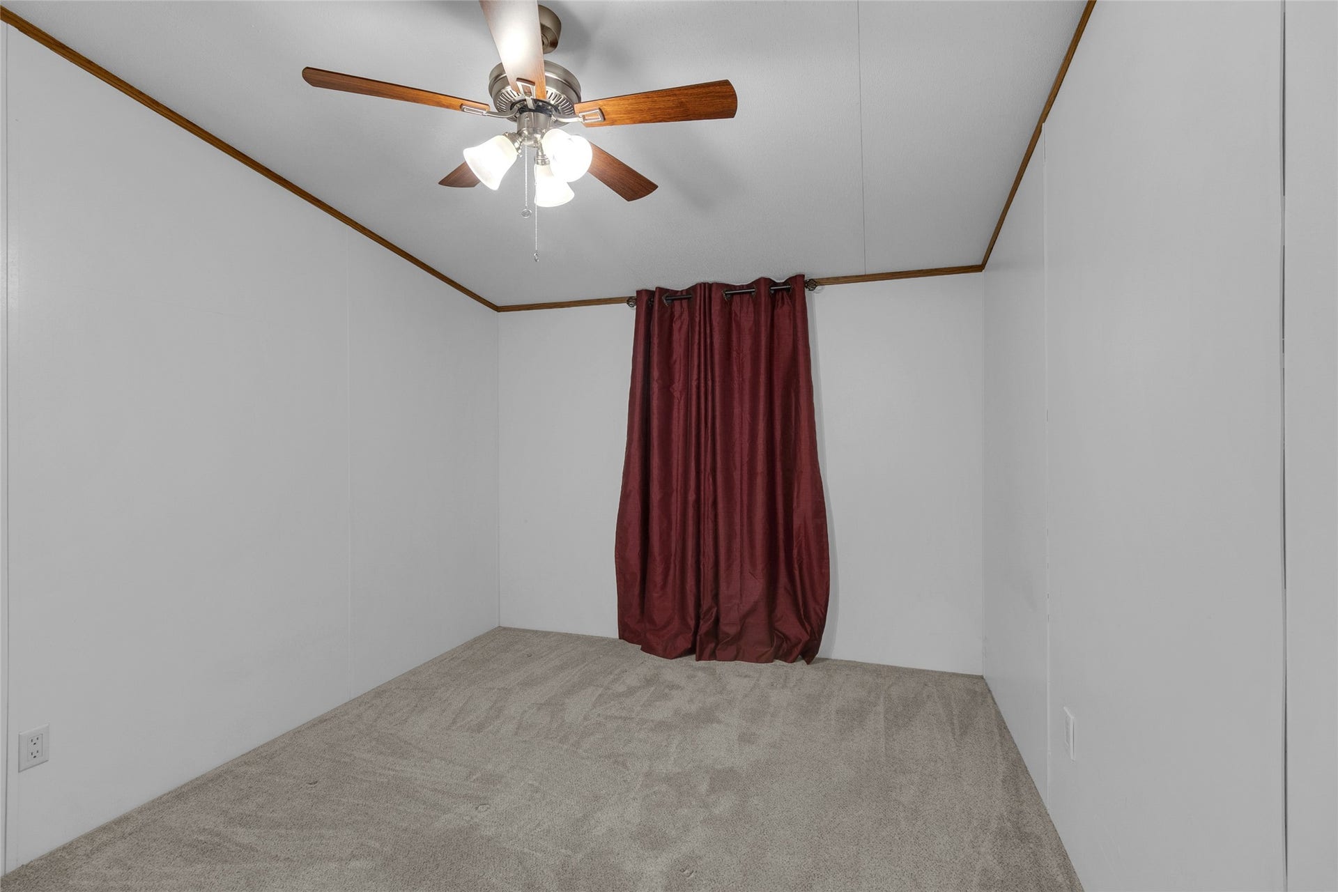 Third bedroom features neutral carpet and walls, accented by a ceiling fan with wooden blades. A window with dark red curtains. The space is simple and versatile, ideal for use as a bedroom or office.