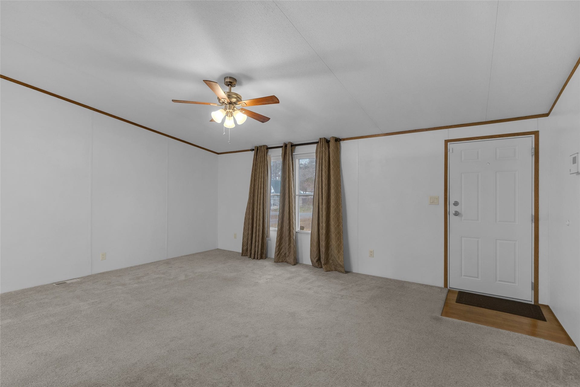 A cozy living room with neutral carpet, a ceiling fan, and a large window with curtains. The space features a simple entry door and clean white walls, offering a bright and inviting atmosphere.