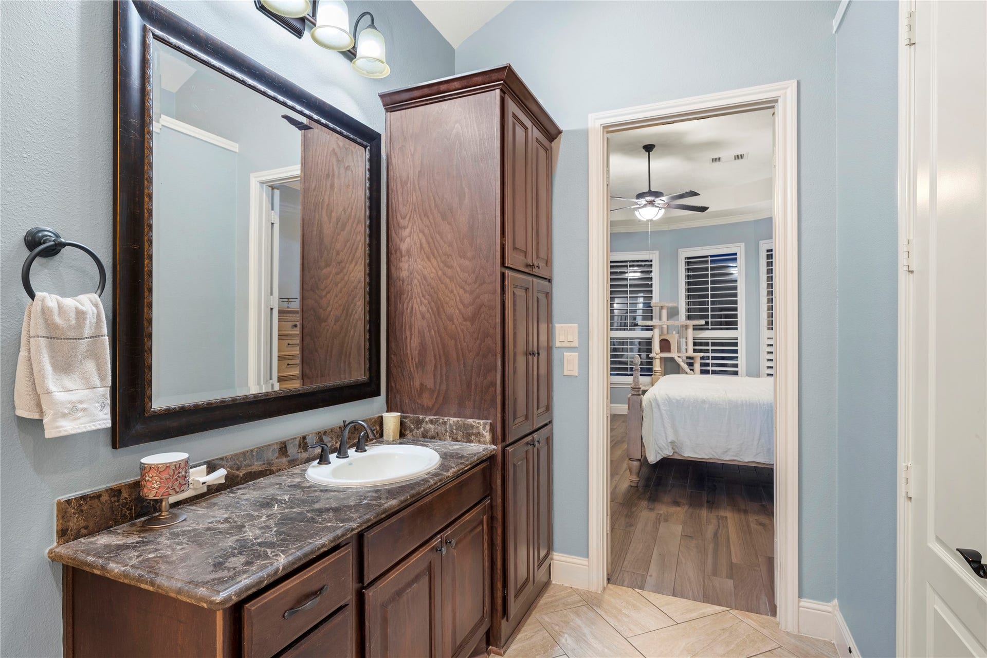 Elegant bathroom featuring rich wood cabinetry, sleek granite countertops, and a large framed mirror, seamlessly transitioning into a cozy bedroom with Italian tile floors and ample natural light.