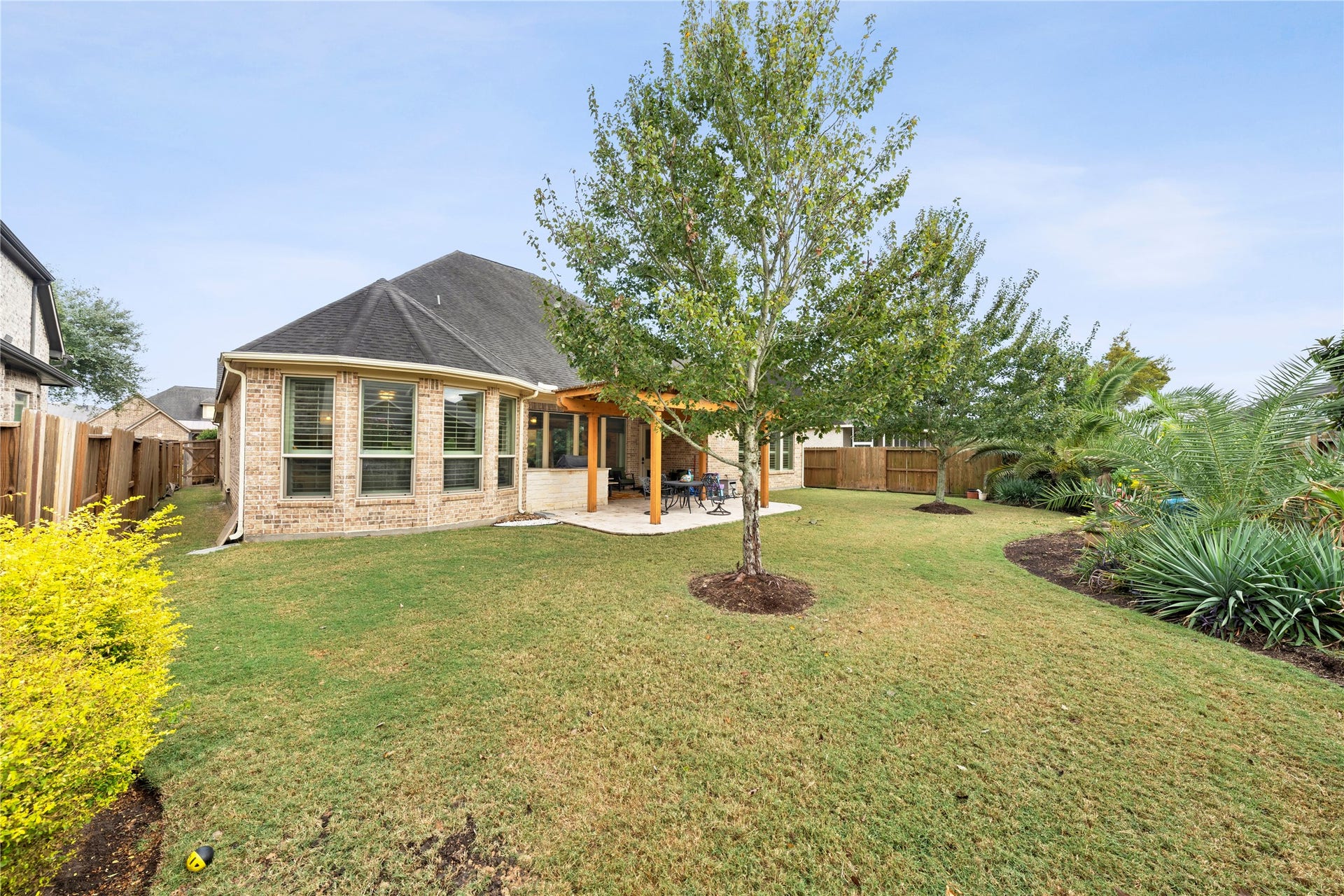 Spacious backyard with lush lawn, mature trees, and a charming brick exterior featuring a covered patio perfect for outdoor gatherings.