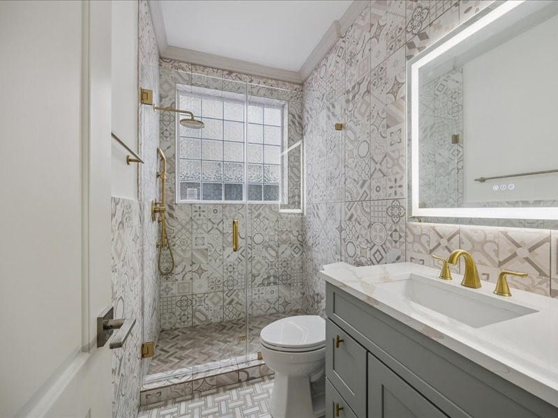 A standout feature of the home, this beautifully renovated bathroom offers a spa-like feel with an open shower, European-style tile throughout, and a sleek backlit mirror. A large frosted window welcomes natural light while preserving privacy.