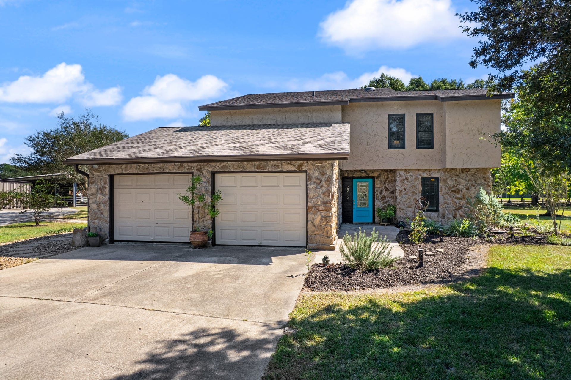 2215 Avenue Q, Santa Fe, TX 77510 Welcoming exterior view of this distinctive two-story home, featuring a beautiful combination of natural stone and stucco, an attached two-car garage, and a charming turquoise front door. Mature shade trees, landscaped garden beds, and a spacious concrete driveway enhance the home's curb appeal, creating an inviting entrance to this unique country property.