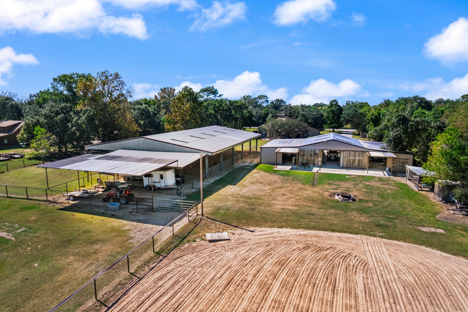 2215 Avenue Q, Santa Fe, TX 77510 Aerial view highlighting the property's impressive equestrian facilities, including a spacious covered horse barn, fenced paddocks, covered trailer parking, and a large sand riding arena. An additional outbuilding provides versatile space for equipment, vehicles, or storage. Thoughtfully arranged for horse care, riding, and everyday ranch activities, this property offers an appealing setup for equestrian enthusiasts.