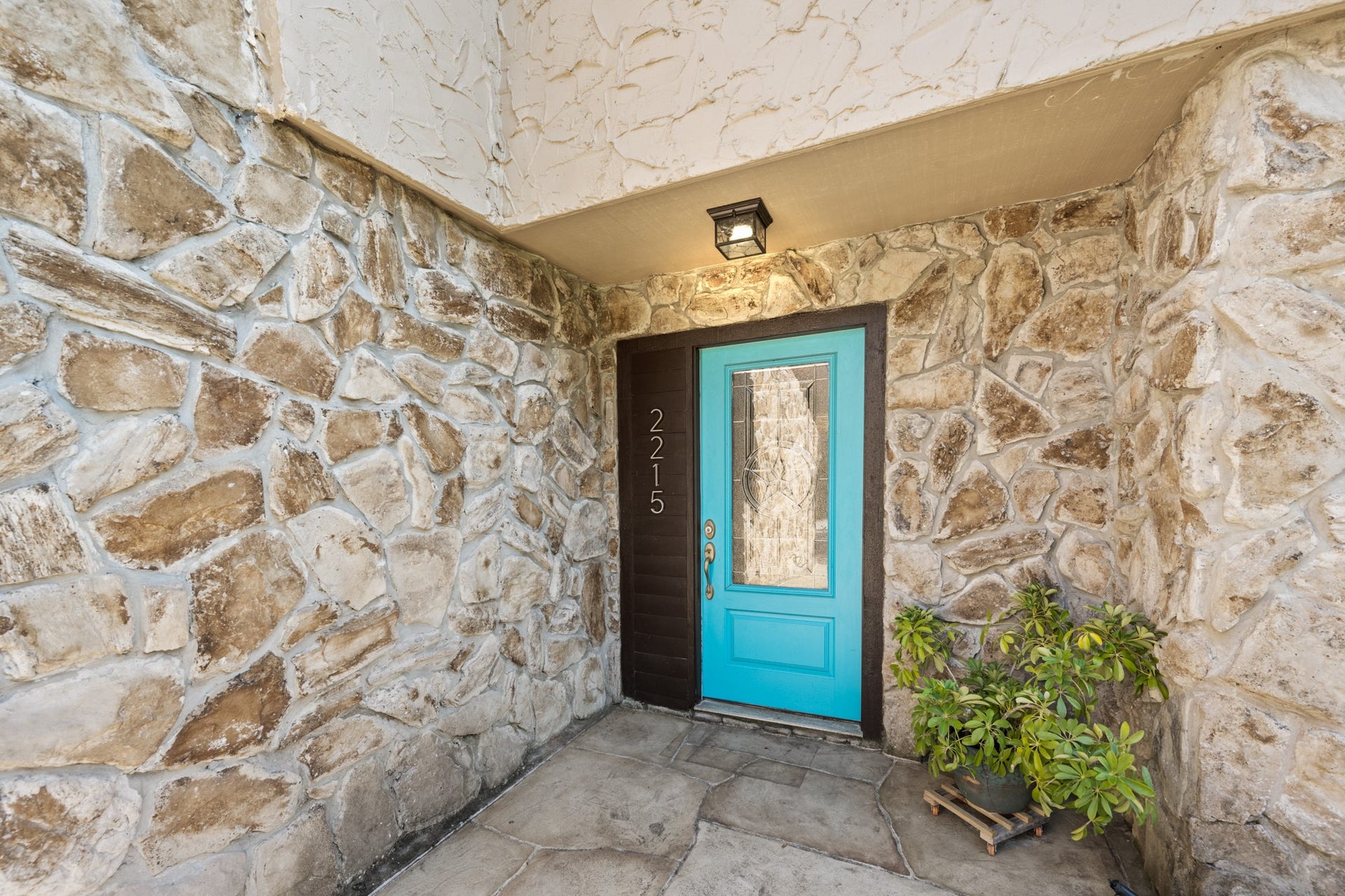 2215 Avenue Q, Santa Fe, TX 77510 A welcoming covered entry showcases rustic stone walls, stone-look flooring, and a striking turquoise front door with decorative glass, adding a vibrant touch of character to the home's exterior.