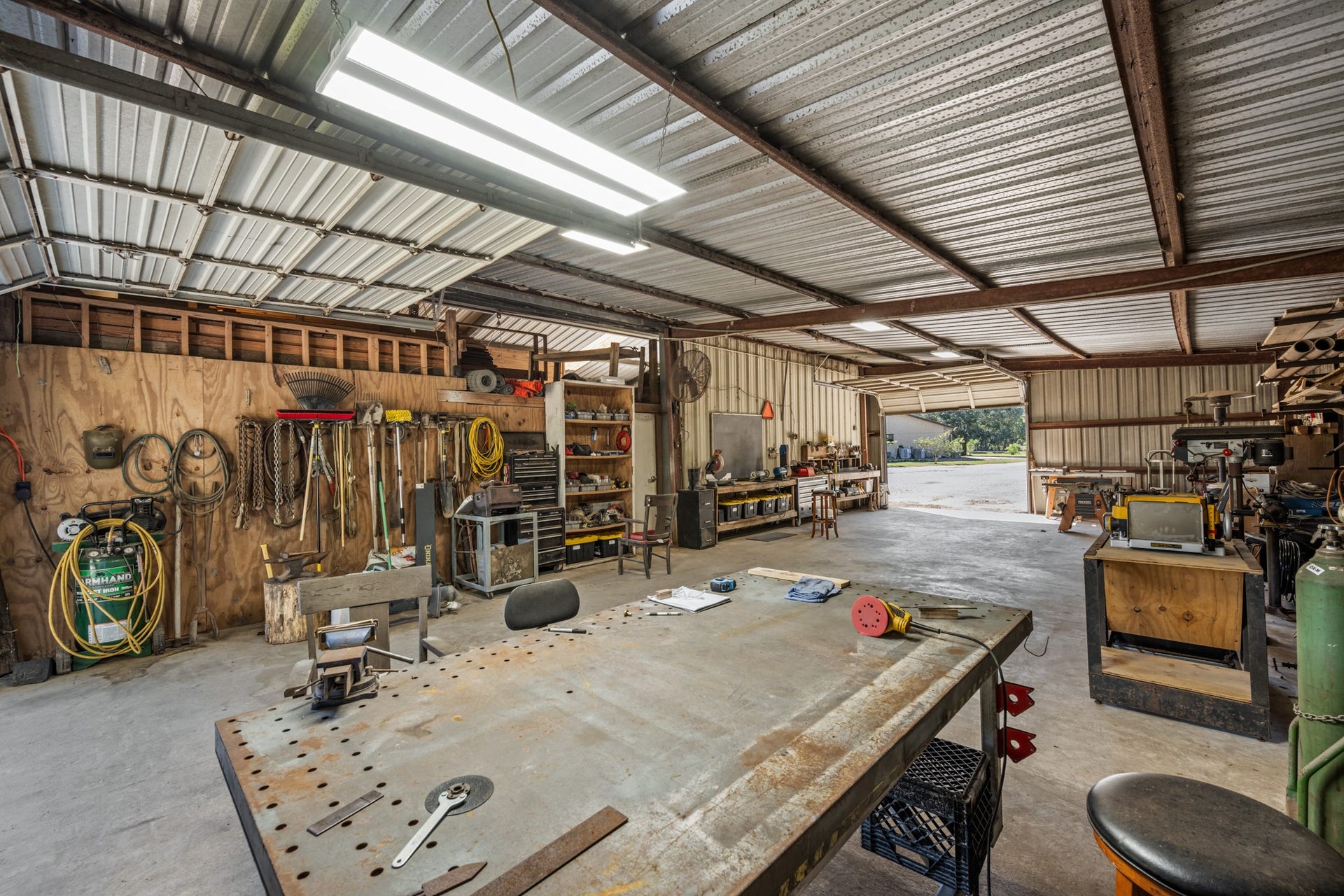2215 Avenue Q, Santa Fe, TX 77510 Another view of the impressive workshop, showcasing a large central worktable, extensive tool storage, built-in workstations, and convenient overhead roll-up door access.