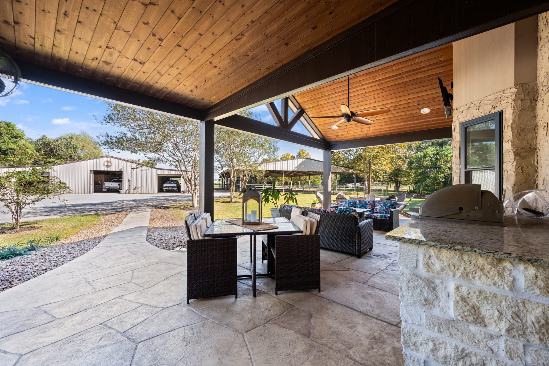 2215 Avenue Q, Santa Fe, TX 77510 A second perspective highlights the covered outdoor kitchen with built-in stainless steel grill, stone-accented countertops, dining space, and comfortable lounge seating. The wood-plank ceiling and exposed timber beams add rustic elegance, with convenient access to the property's spacious outbuildings.