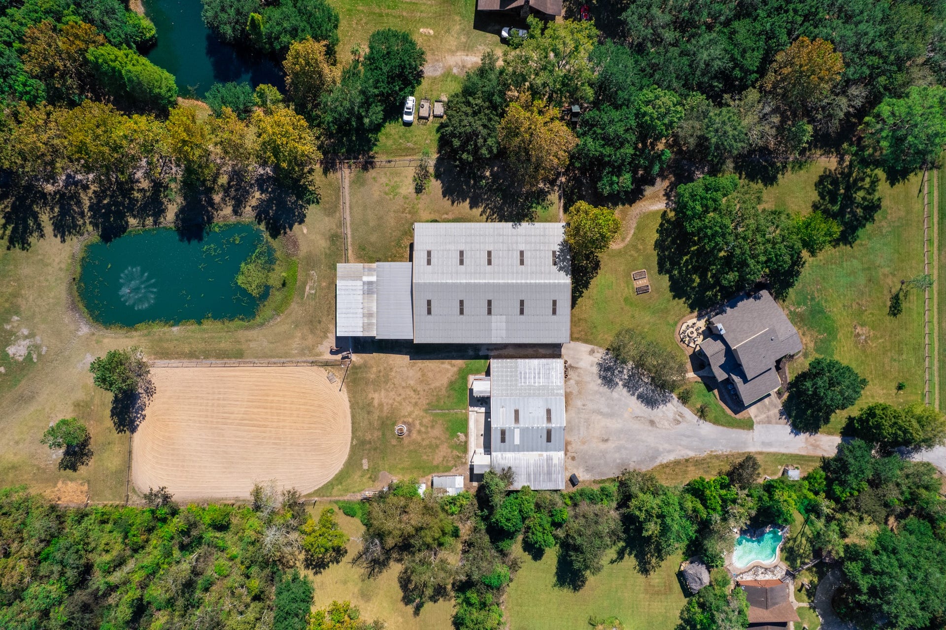 2215 Avenue Q, Santa Fe, TX 77510 Overhead aerial view showcasing the property's impressive layout, featuring a spacious horse barn, sand riding arena, scenic pond with fountain, additional outbuilding, and residence surrounded by mature trees. The thoughtfully arranged equestrian facilities offer convenient access between the barn, arena, and surrounding grounds, creating an ideal setting for horse enthusiasts seeking space, functionality, and country living.