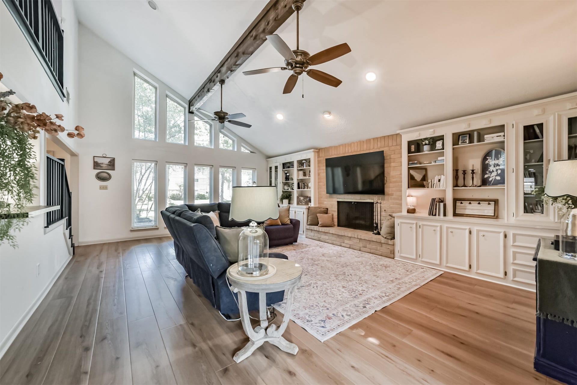 7618 Hurst Forest Drive, Humble, TX 77346 A brick fireplace anchors the living room, creating a cozy gathering spot beneath the dramatic vaulted ceiling. Built-in cabinetry combines useful storage with display space, and tall windows fill the room with natural light.