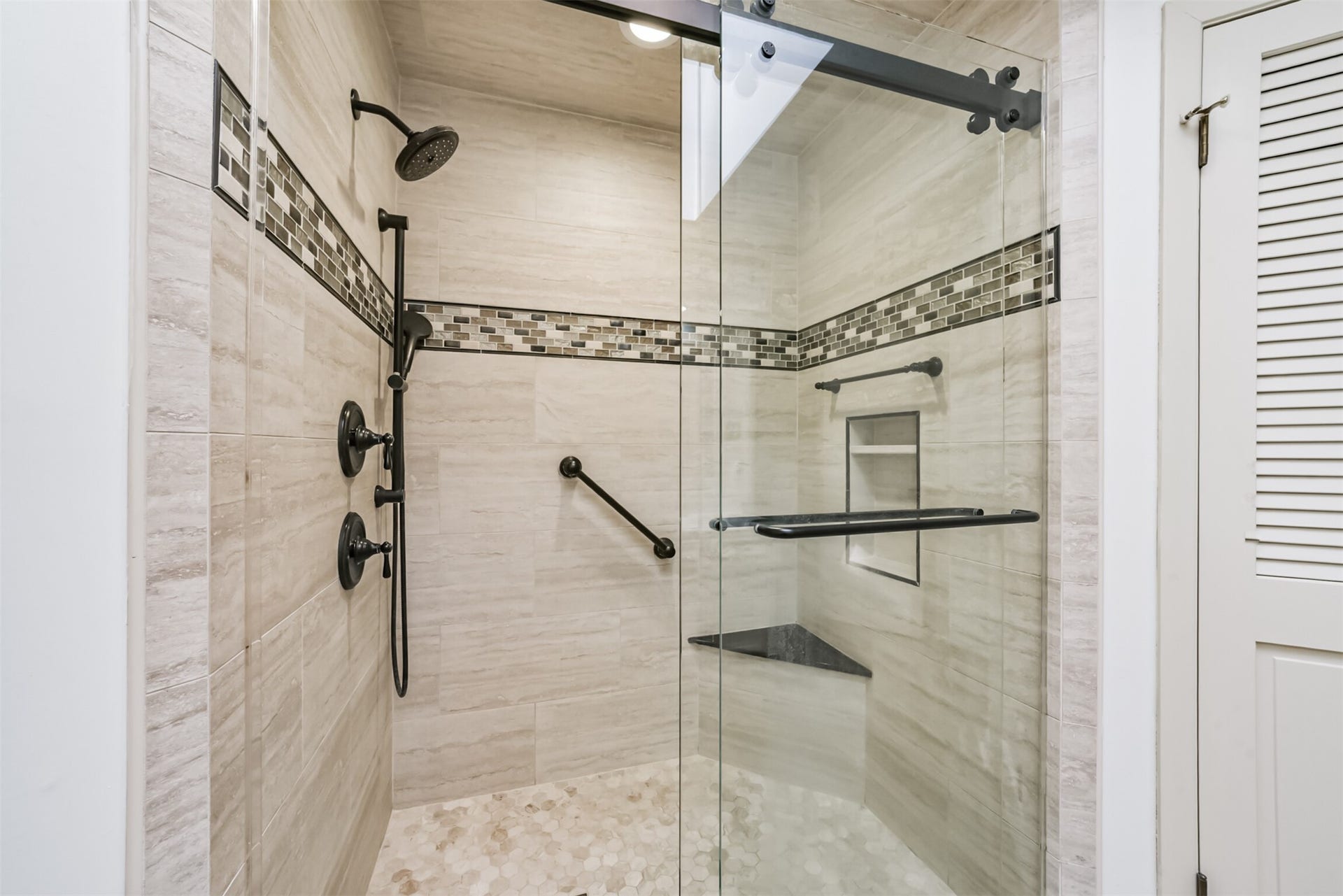 7618 Hurst Forest Drive, Humble, TX 77346 The spacious walk-in shower pairs warm tile with contrasting fixtures and a sliding glass enclosure. A built-in bench, recessed niche, and handheld shower add thoughtful convenience to this relaxing space.