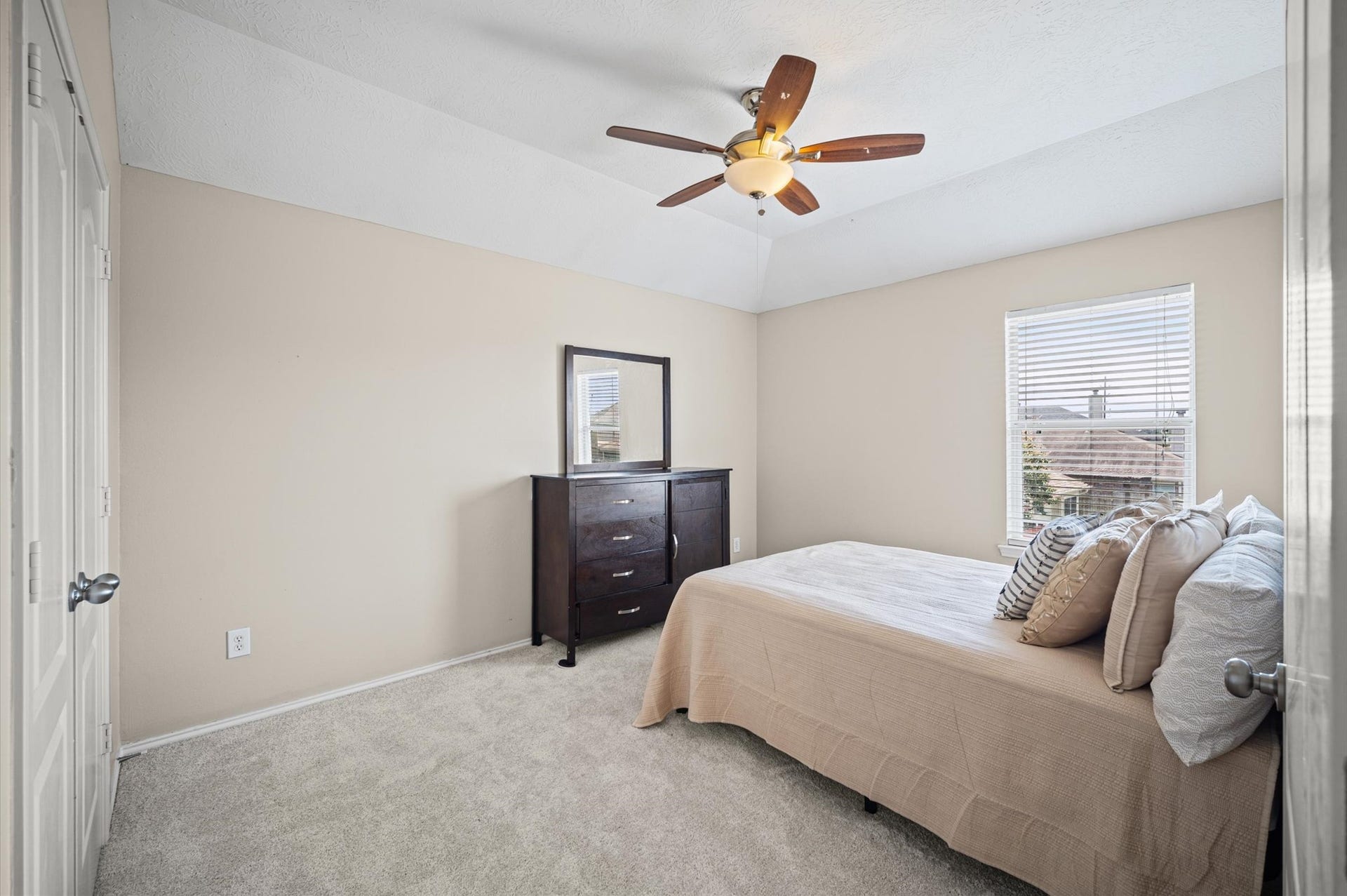 8711 Sandusky Court, Tomball, TX 77375 This cozy bedroom features neutral walls, a ceiling fan, and a single window for natural light. It includes a comfortable bed and a dark wood dresser with a mirror, offering a simple and inviting layout.