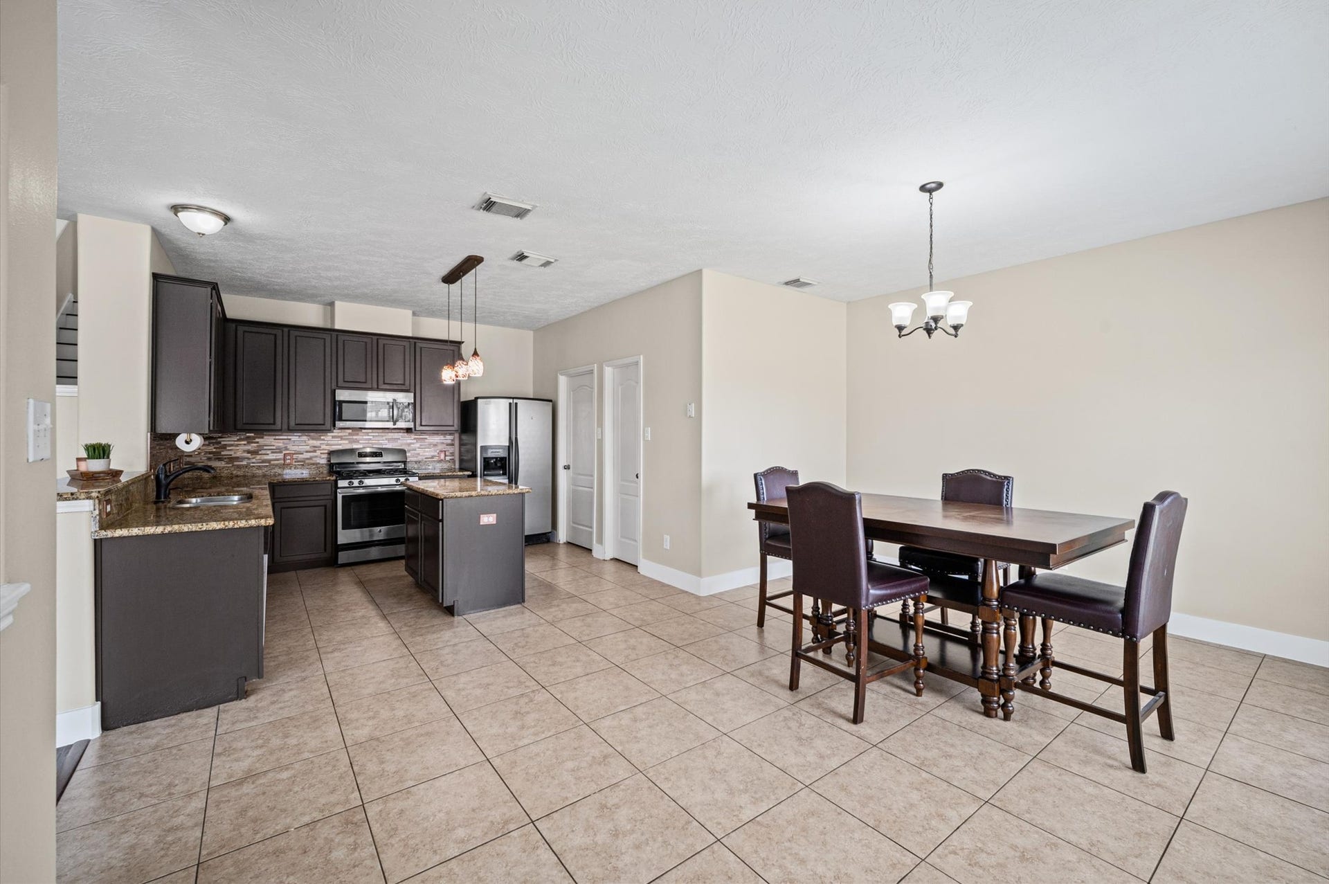8711 Sandusky Court, Tomball, TX 77375 This photo showcases a spacious open-concept kitchen and dining area. The kitchen features dark wood cabinets, granite countertops, and stainless steel appliances with a tile backsplash. The adjacent dining area has a modern chandelier and ample space for a table, all set on neutral tile flooring.