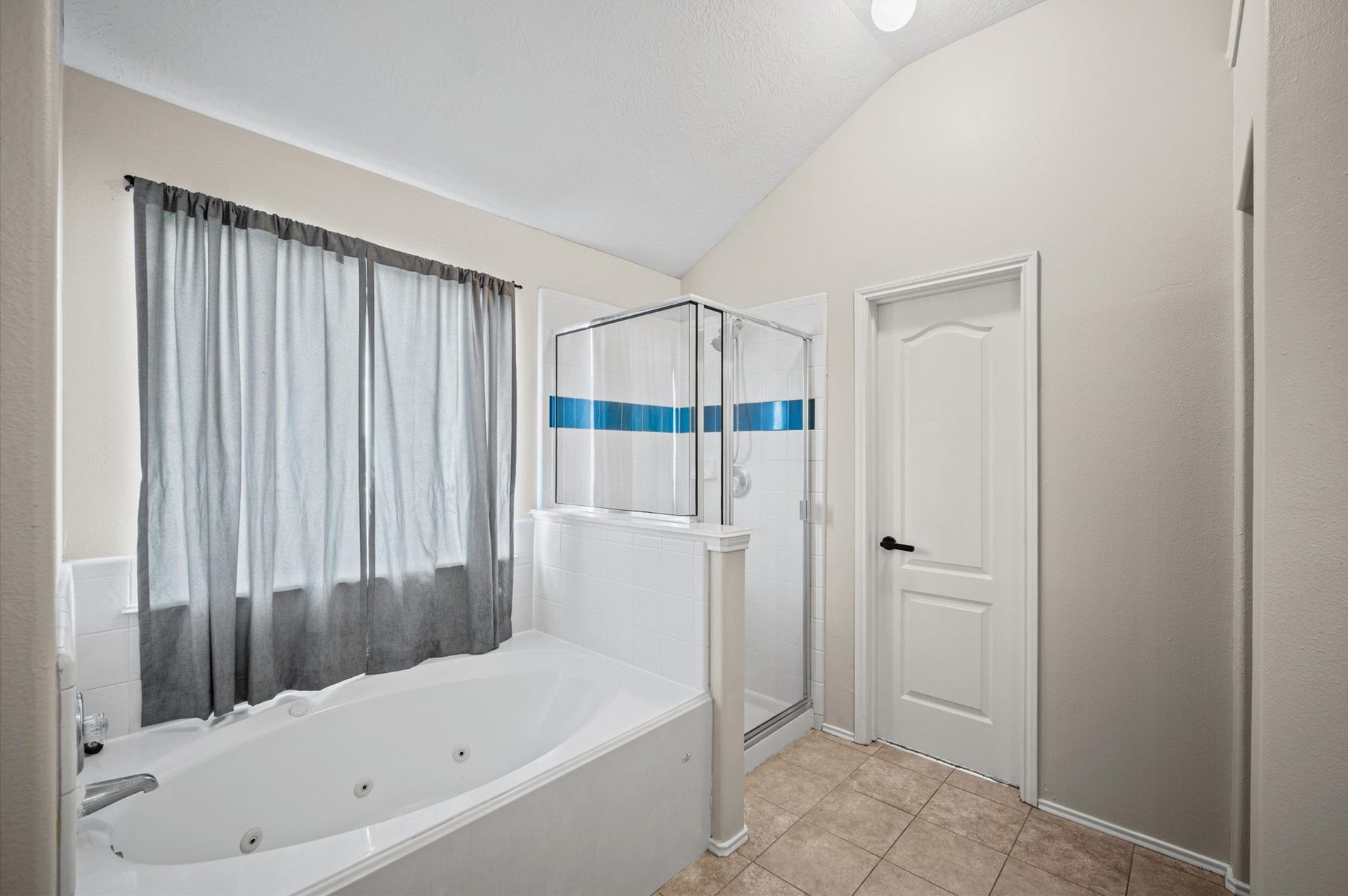 8711 Sandusky Court, Tomball, TX 77375 This bathroom features a spacious white jetted tub with a curtain, a separate glass-enclosed shower with blue accent tiles, and a white door. It has a neutral color palette and a tiled floor.