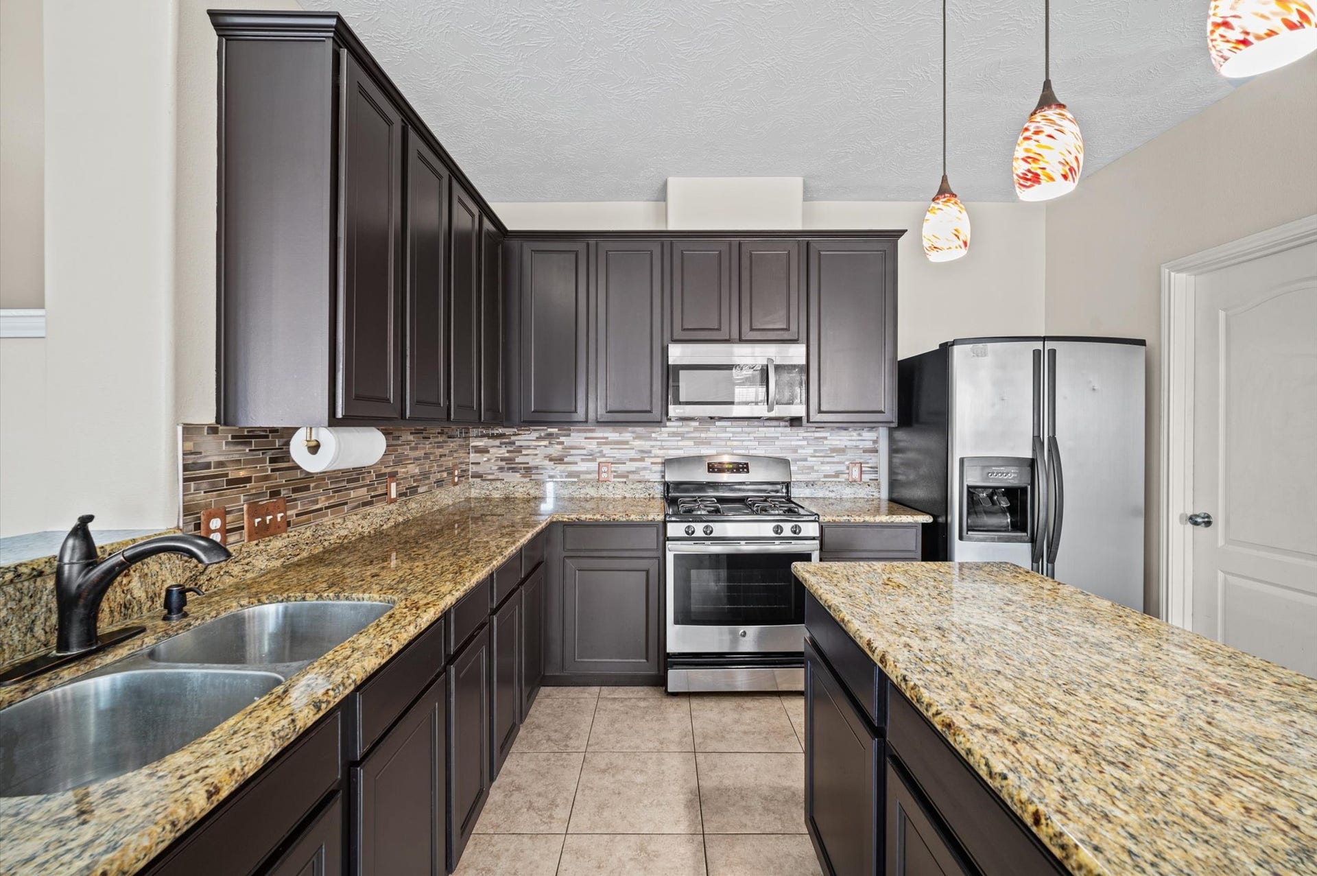 8711 Sandusky Court, Tomball, TX 77375 Modern kitchen with dark cabinetry, granite countertops, and stainless steel appliances. Features a stylish tile backsplash, double sink, and pendant lighting for a sleek, contemporary look.