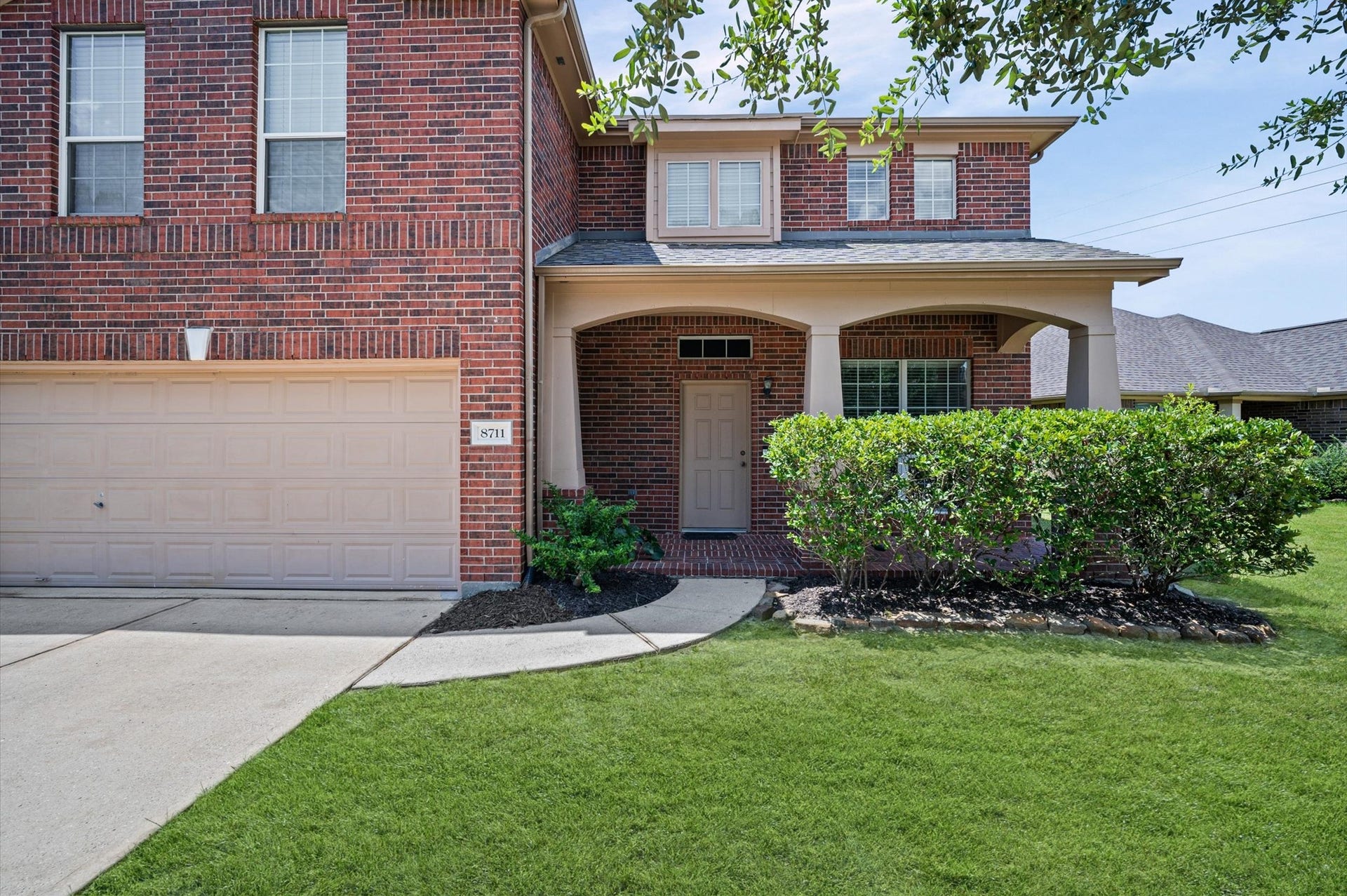 8711 Sandusky Court, Tomball, TX 77375 This photo shows a two-story brick home with a welcoming front porch and attached two-car garage. The well-maintained lawn and shrubs enhance its curb appeal, while the covered entry provides a sheltered welcome.