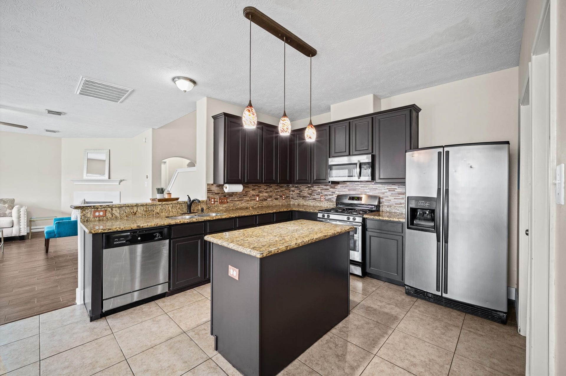 8711 Sandusky Court, Tomball, TX 77375 This modern kitchen features dark cabinetry, granite countertops, and stainless steel appliances. It includes a center island with pendant lighting and opens to a spacious living area with tiled flooring.