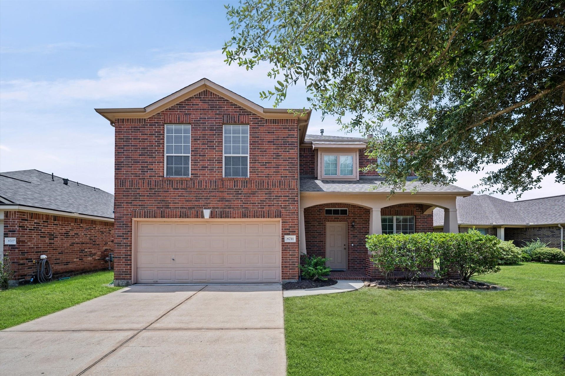 8711 Sandusky Court, Tomball, TX 77375 This photo shows a charming two-story brick home with a two-car garage and a covered front porch. It features well-maintained landscaping with lush grass and bushes, situated in a suburban neighborhood setting.
