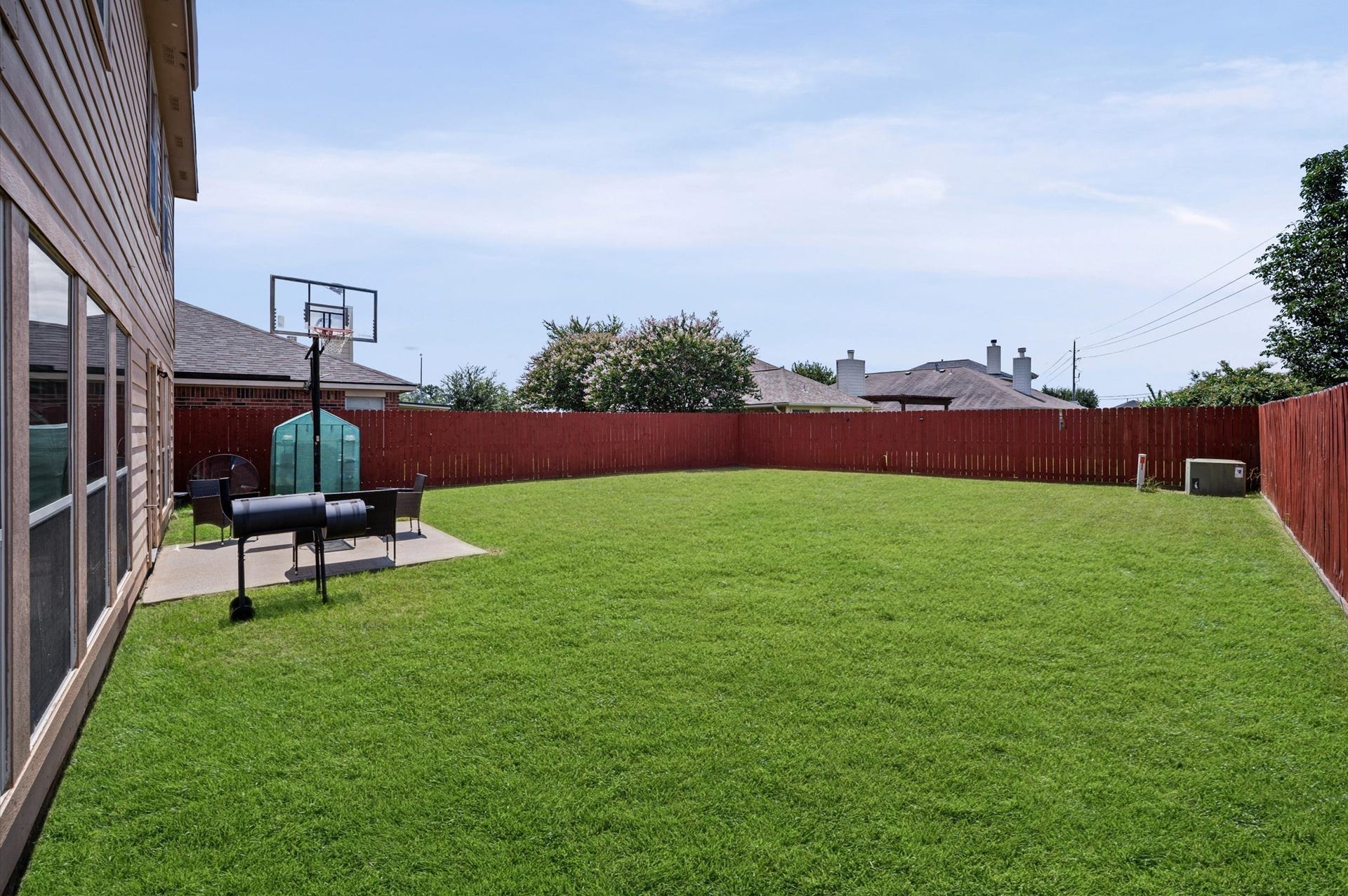 8711 Sandusky Court, Tomball, TX 77375 Spacious backyard with a well-maintained lawn, red wooden fence, and a concrete patio area featuring a grill and basketball hoop. Ideal for outdoor activities and entertaining.