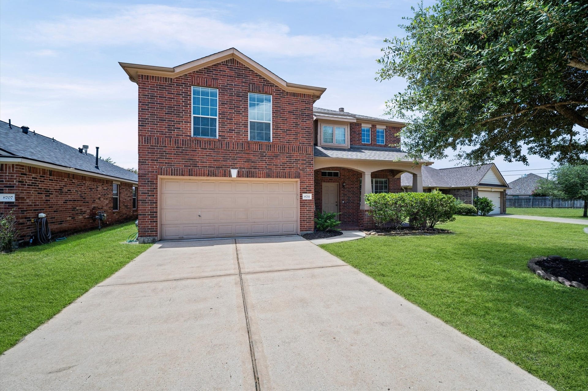 8711 Sandusky Court, Tomball, TX 77375 This photo shows a two-story brick house with a two-car garage and a neatly maintained front yard. The driveway is spacious, and the house features multiple large windows. There's a small garden area with shrubs near the entrance, and the neighborhood appears quiet and well-kept.