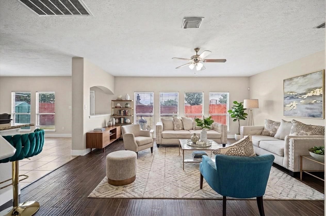 8711 Sandusky Court, Tomball, TX 77375 This spacious living room features elegant wood flooring, ample natural light from large windows, and comfortable seating with stylish, neutral-toned furniture. A ceiling fan and an adjacent open dining area enhance the inviting atmosphere.