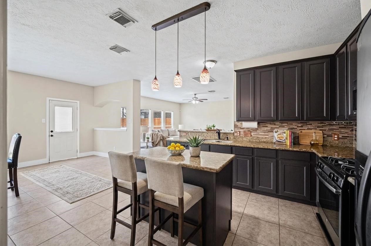 8711 Sandusky Court, Tomball, TX 77375 This spacious kitchen features dark wood cabinets, granite countertops, and a central island with pendant lighting. It opens to a bright living area with large windows and tile flooring, offering a modern and inviting space.