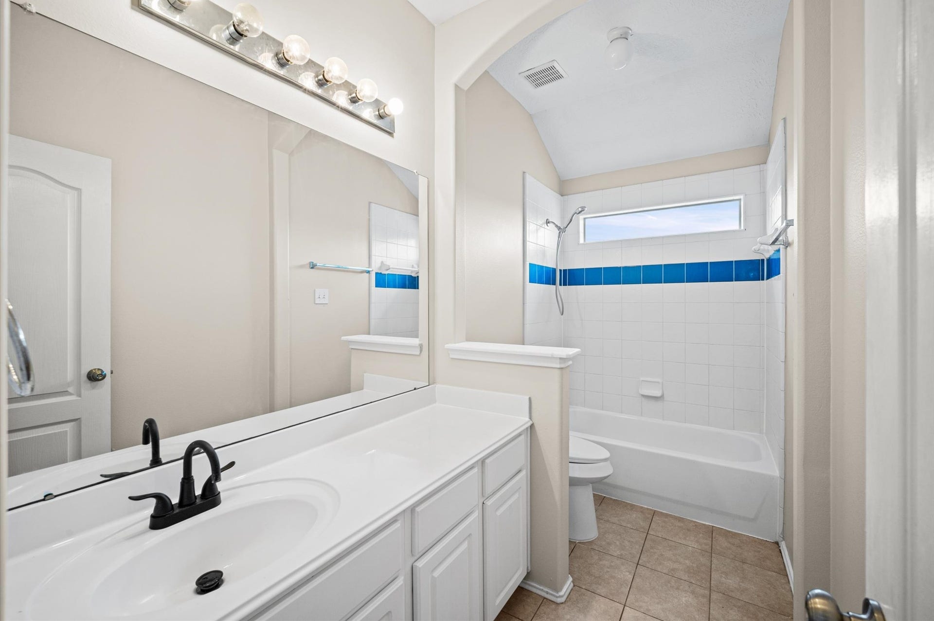 8711 Sandusky Court, Tomball, TX 77375 This bathroom features a clean, modern design with a large mirror and vanity, a bathtub with a shower, and bright lighting. The white tiles are accented with a bold blue stripe, adding a touch of color.