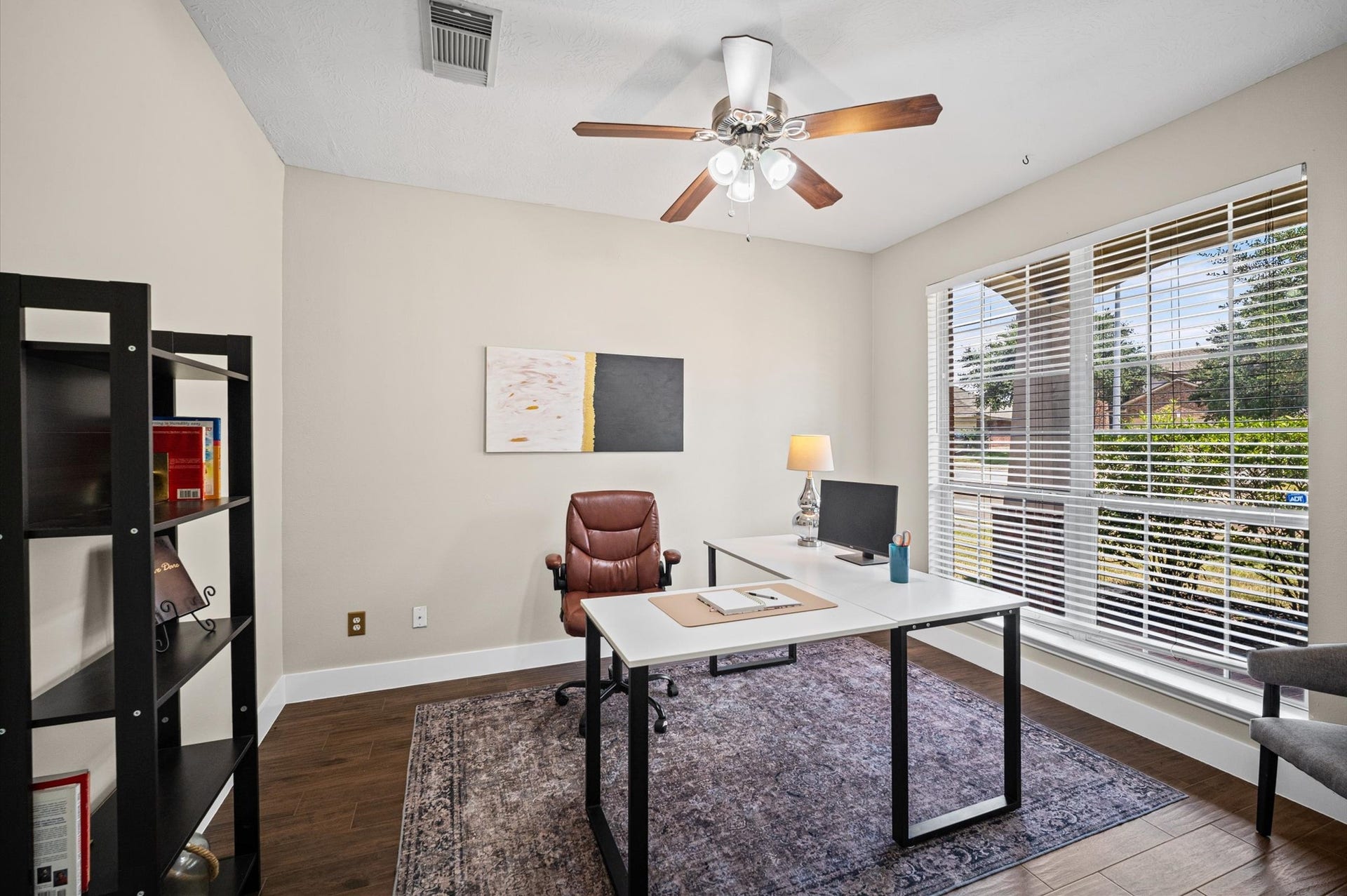 8711 Sandusky Court, Tomball, TX 77375 Bright home office with large windows, modern ceiling fan, and hardwood floors. Features a sleek desk, leather chair, and a bookshelf. Ideal for productivity with natural light and a view of the outdoors.