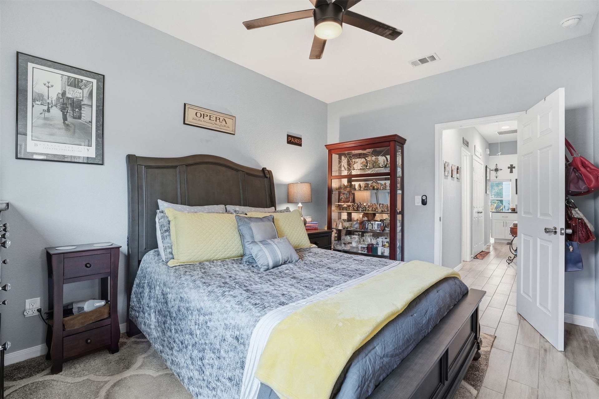 16740 Stonefield, Montgomery, TX 77316 Primary bedroom with a stylish dark wood bed frame, complemented by soft gray walls and bright yellow accents. A glass display cabinet adds charm, while the ceiling fan ensures comfort. The room connects to a bathroom, enhancing convenience.