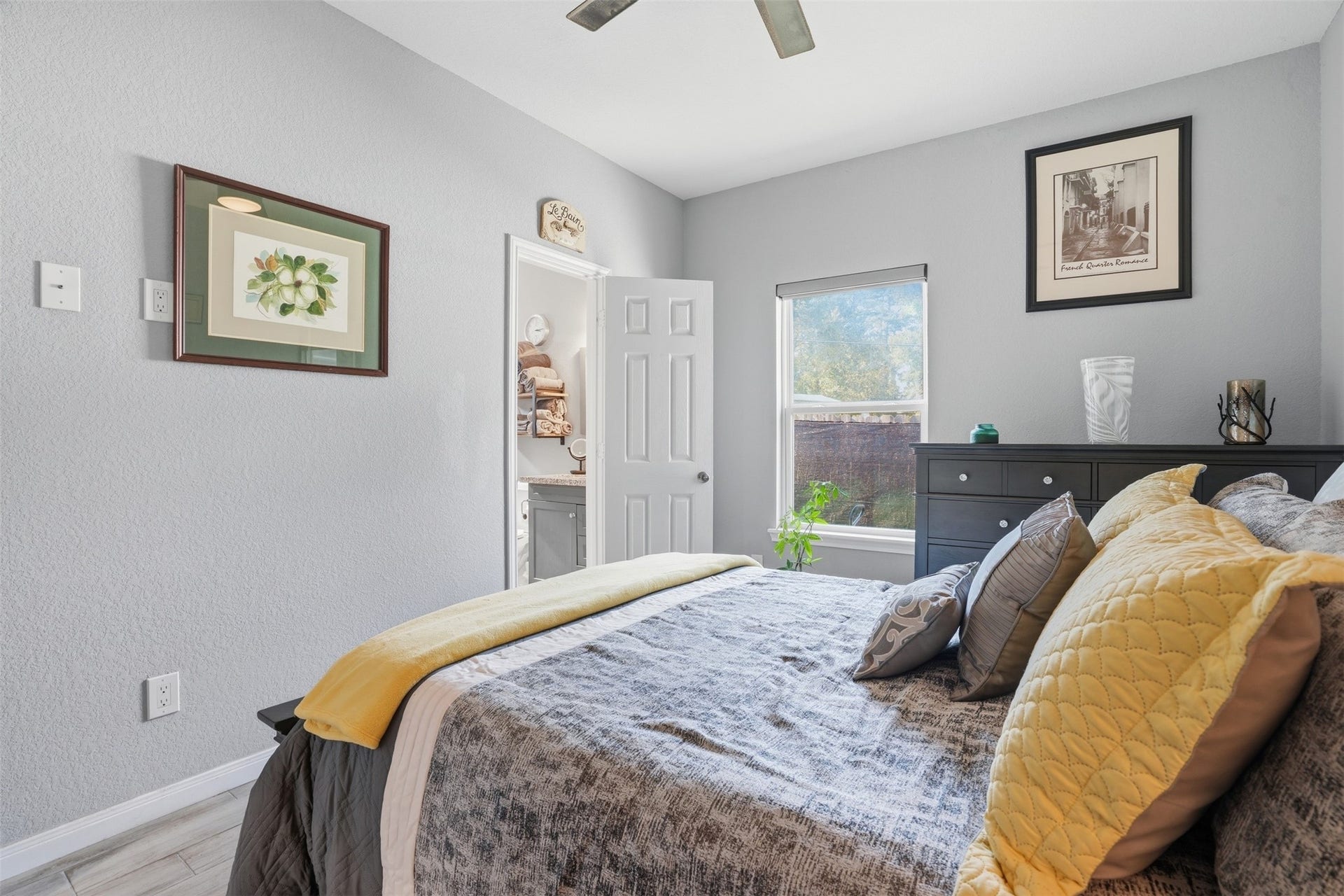 16740 Stonefield, Montgomery, TX 77316 Primary bedroom with soft gray walls, featuring a neatly made bed adorned with colorful pillows, ample natural light from a large window, and direct access to a en-suite bathroom.