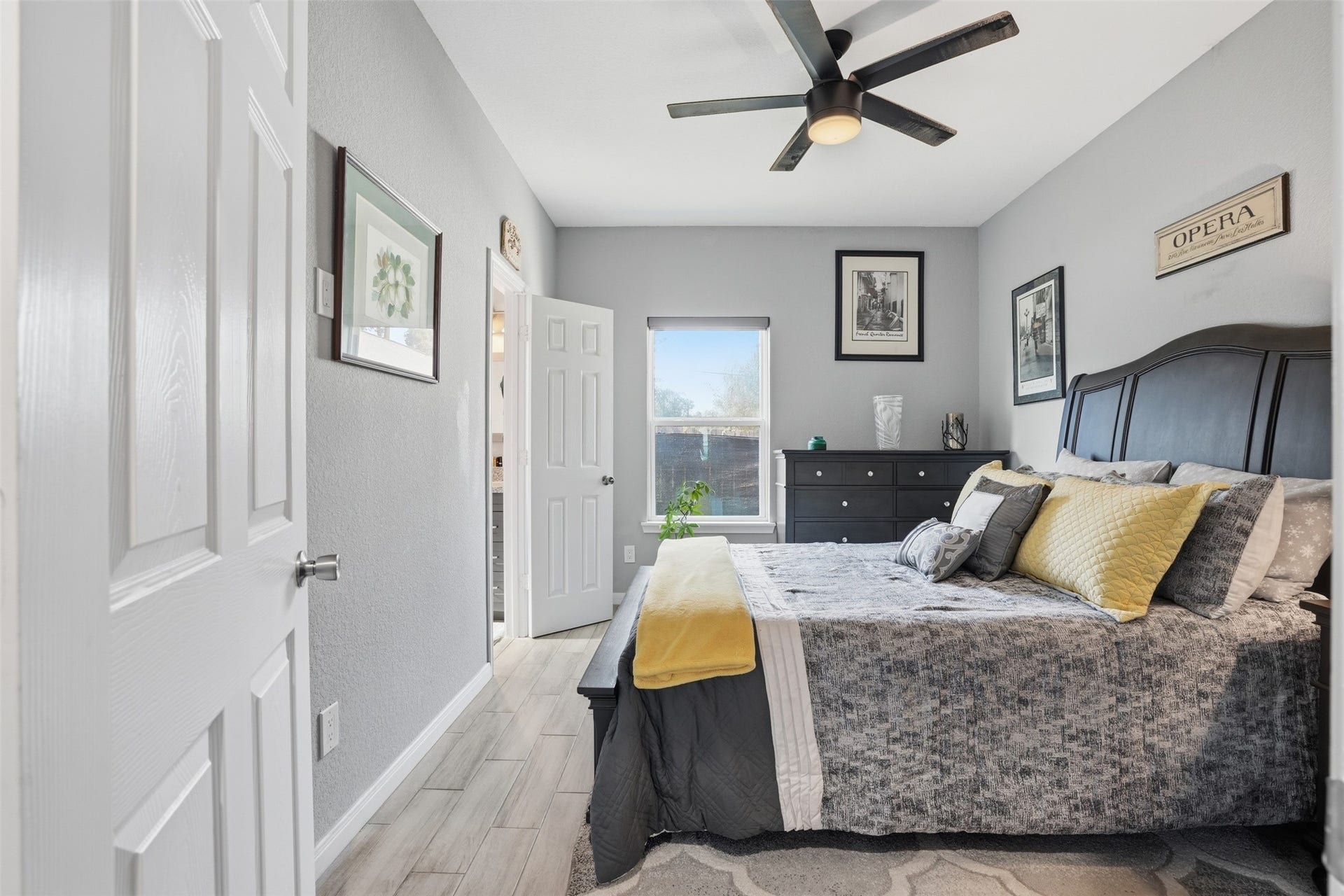 16740 Stonefield, Montgomery, TX 77316 Primary bedroom with soft gray walls, and a large window that allows natural light to fill the space. The room is elegantly decorated with framed artwork and a plush bed adorned with complementary pillows.