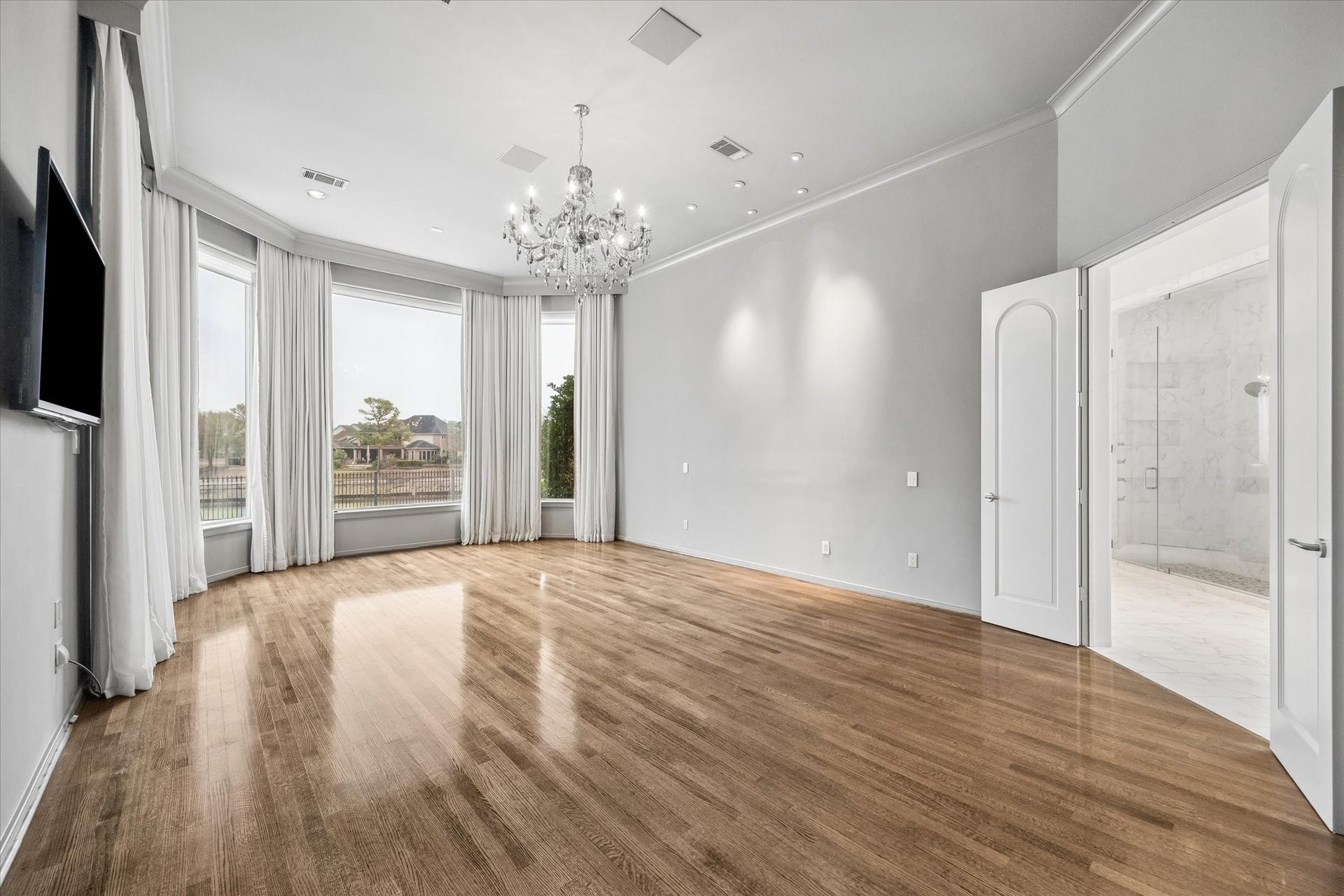 SERENE PRIMARY BEDROOM WITH TALL WINDOWS PROVIDING BEAUTIFUL VIEWS OVER THE LAKE AND GOLF COURSE - pale silvery/gray walls and handsome hardwood flooring, decorative chandelier and recessed lighting, door on left wall leads to the multi-level covered terrace and to the pool/spa