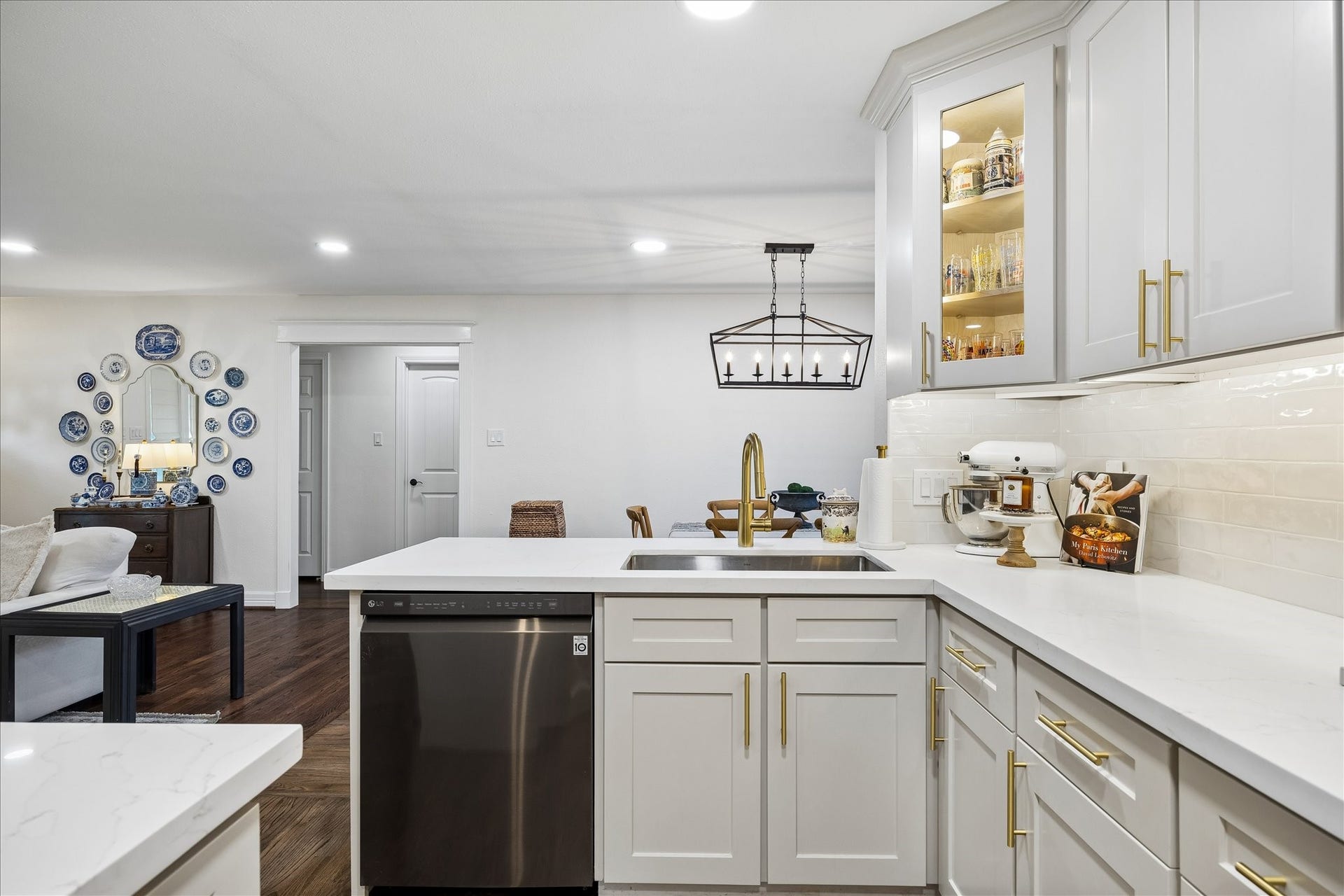 Elegant kitchen with sleek white cabinetry, gold hardware, and modern stainless steel appliances. The open layout flows into a cozy living area featuring rich hardwood floors and tasteful decor accents.