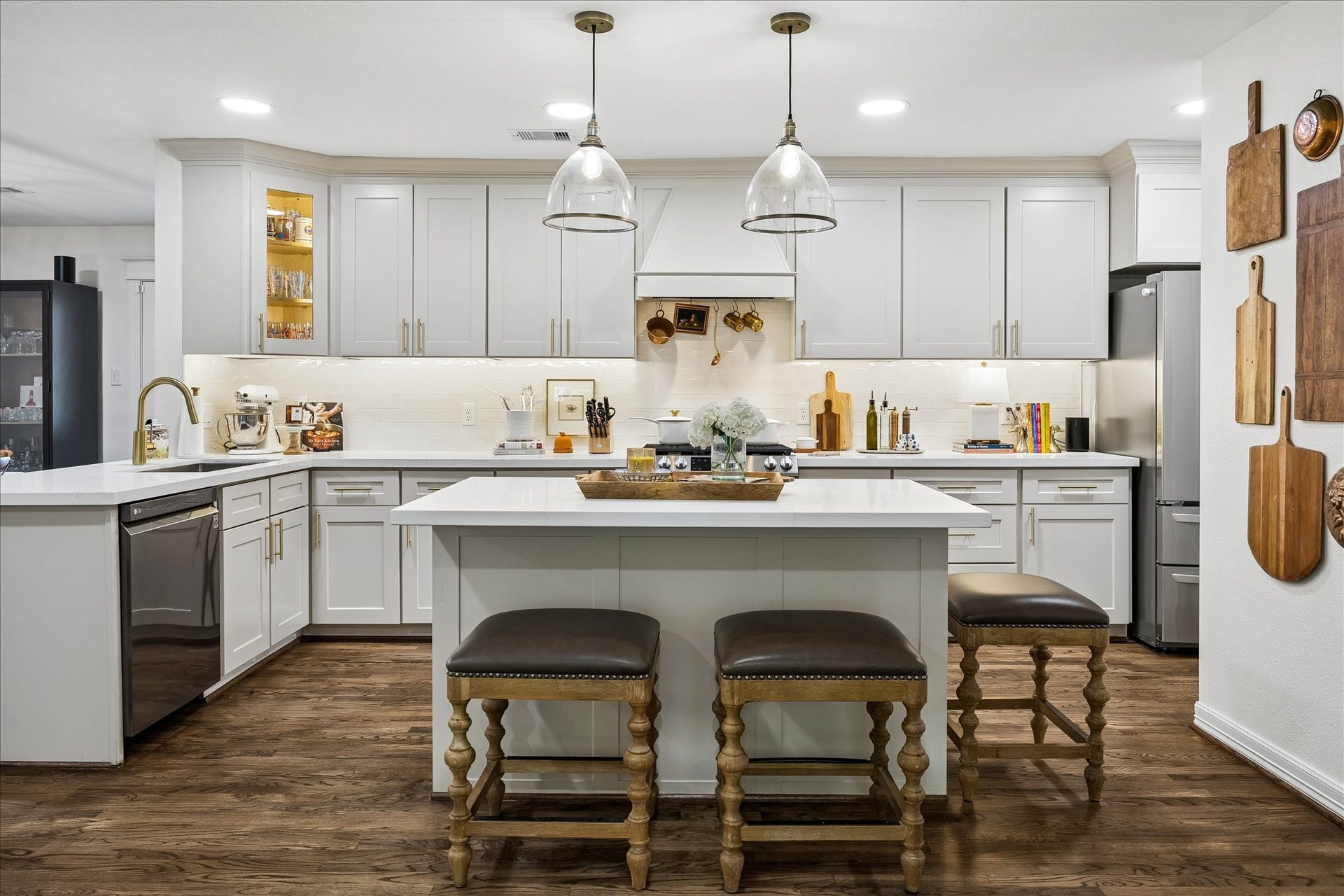 View of kitchen from family room. Sleek white cabinetry, stainless steel appliances, and a central island featuring stylish pendant lighting and seating, perfect for entertaining.