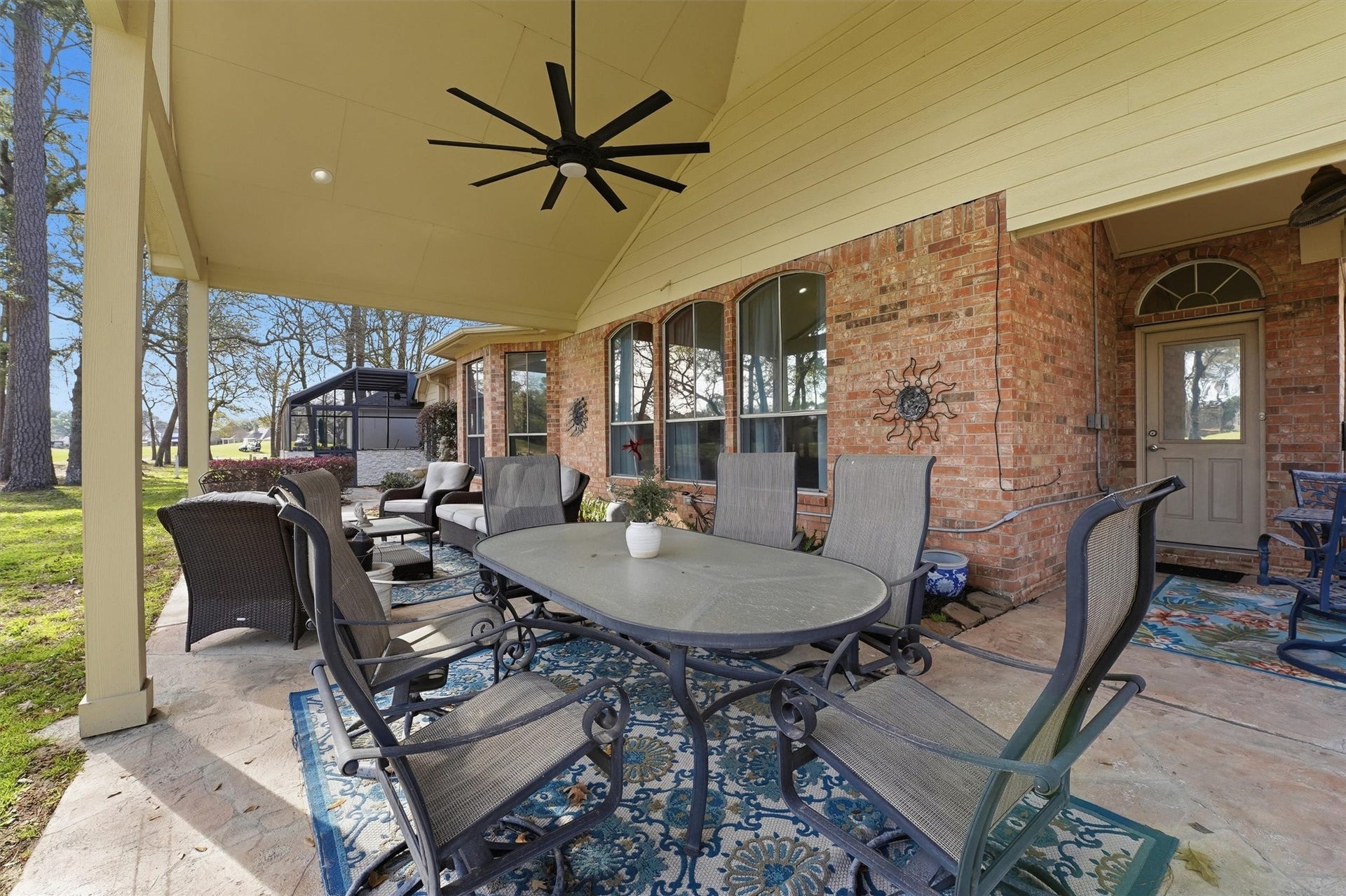 This photo showcases a spacious covered patio with comfortable seating, a dining set, and a ceiling fan. The area features a brick exterior, decorative elements, and overlooks a tree-filled yard, perfect for outdoor entertaining.  This house is located on the golf course, hole 3.