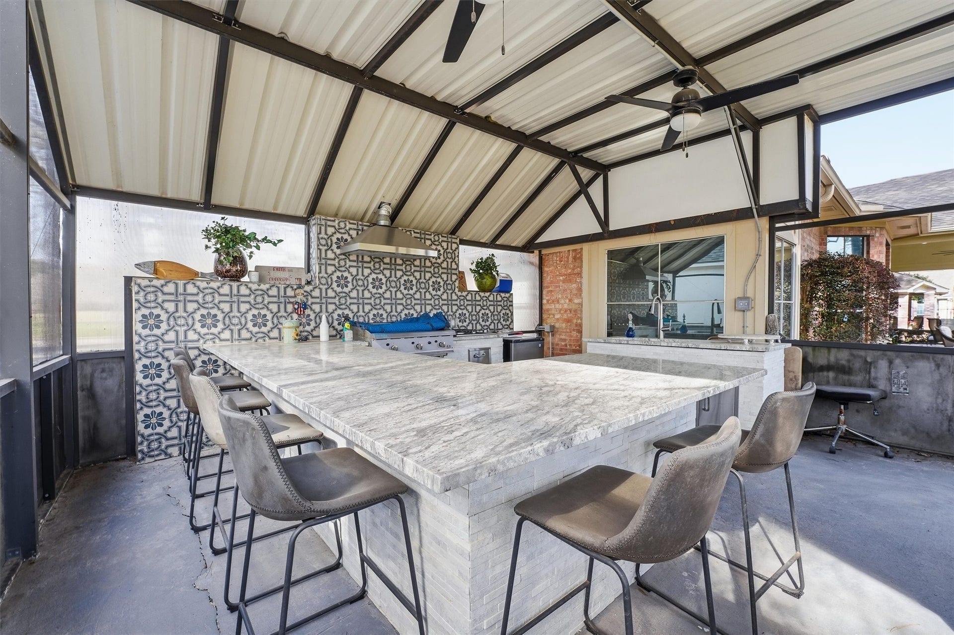 This photo showcases a modern outdoor kitchen with a spacious granite countertop and bar seating. The area features a stylish patterned tile backsplash, built-in grill, and ceiling fans, perfect for entertaining. The covered space offers ample natural light and ventilation.