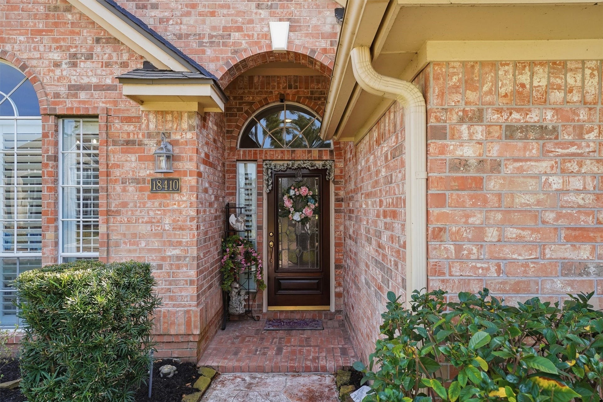 This inviting red brick home features a charming front entry with an arched doorway, decorative glass door, and lush landscaping. A welcoming and well-maintained exterior.