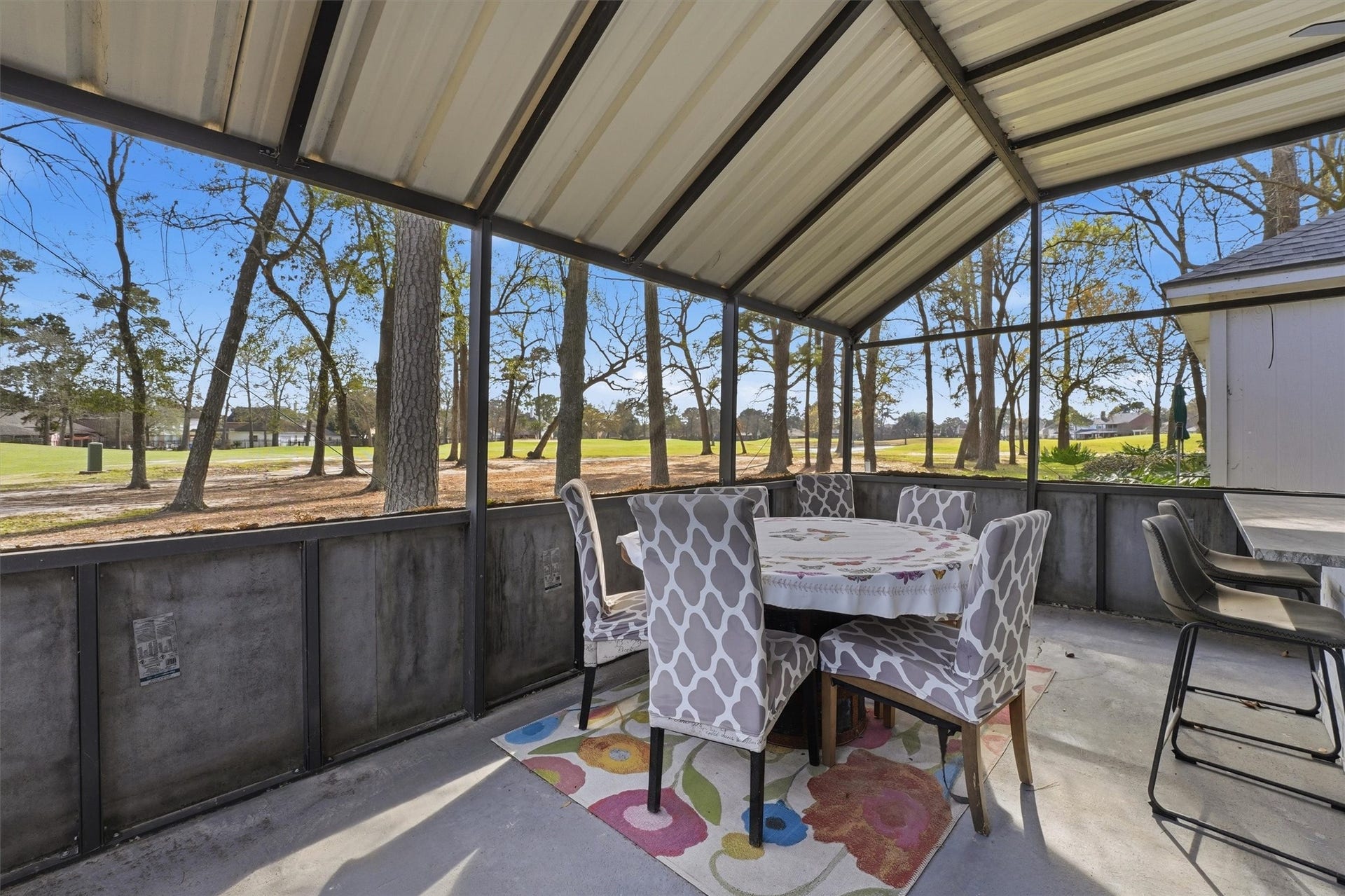 This photo showcases a covered outdoor patio with a dining area, featuring a table and chairs on a decorative rug. The space offers a scenic view of a lush, tree-lined golf course. Ideal for enjoying meals and relaxation in a serene setting.