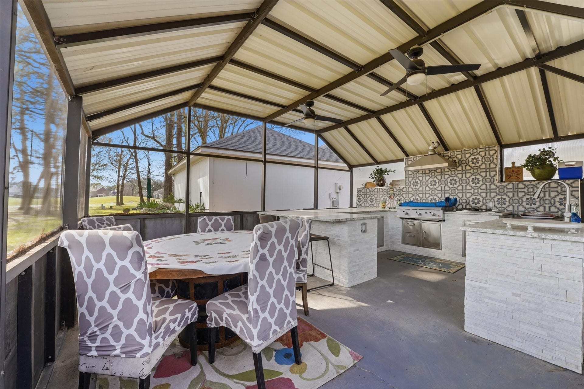 This outdoor living space features a covered patio with a modern kitchen equipped with a large flat top griddle, 4 burner gas cooktop, refrigerator, ceiling fans, stainless steel drawers and cabinets and a sink. The area includes stylish tile work and a dining table with patterned chairs, perfect for entertaining.