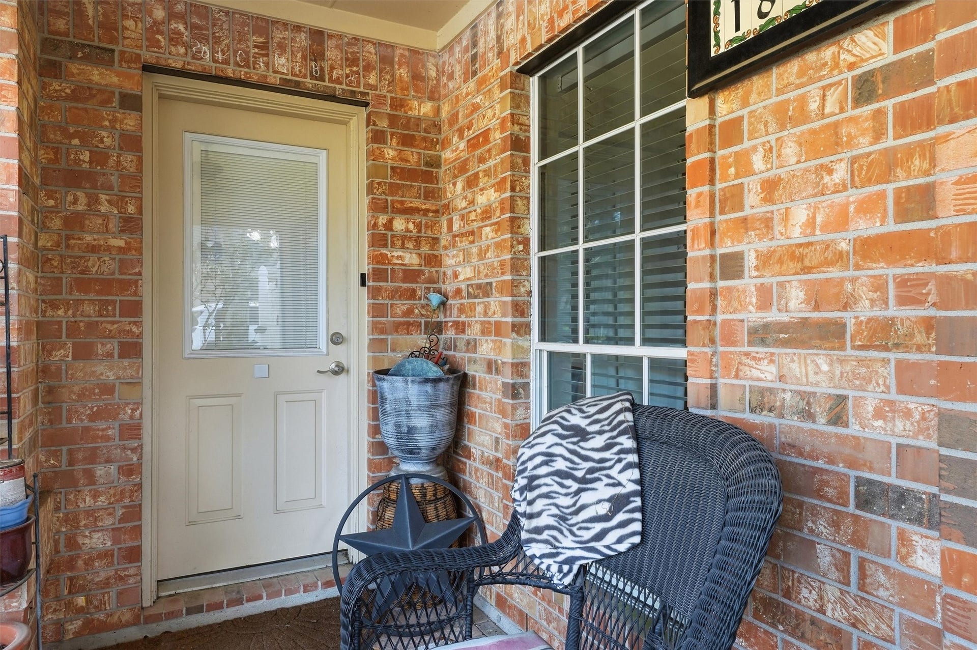 Cozy brick porch with a welcoming door, a large window, and a charming seating area featuring a wicker chair and decorative elements. Perfect for relaxing.