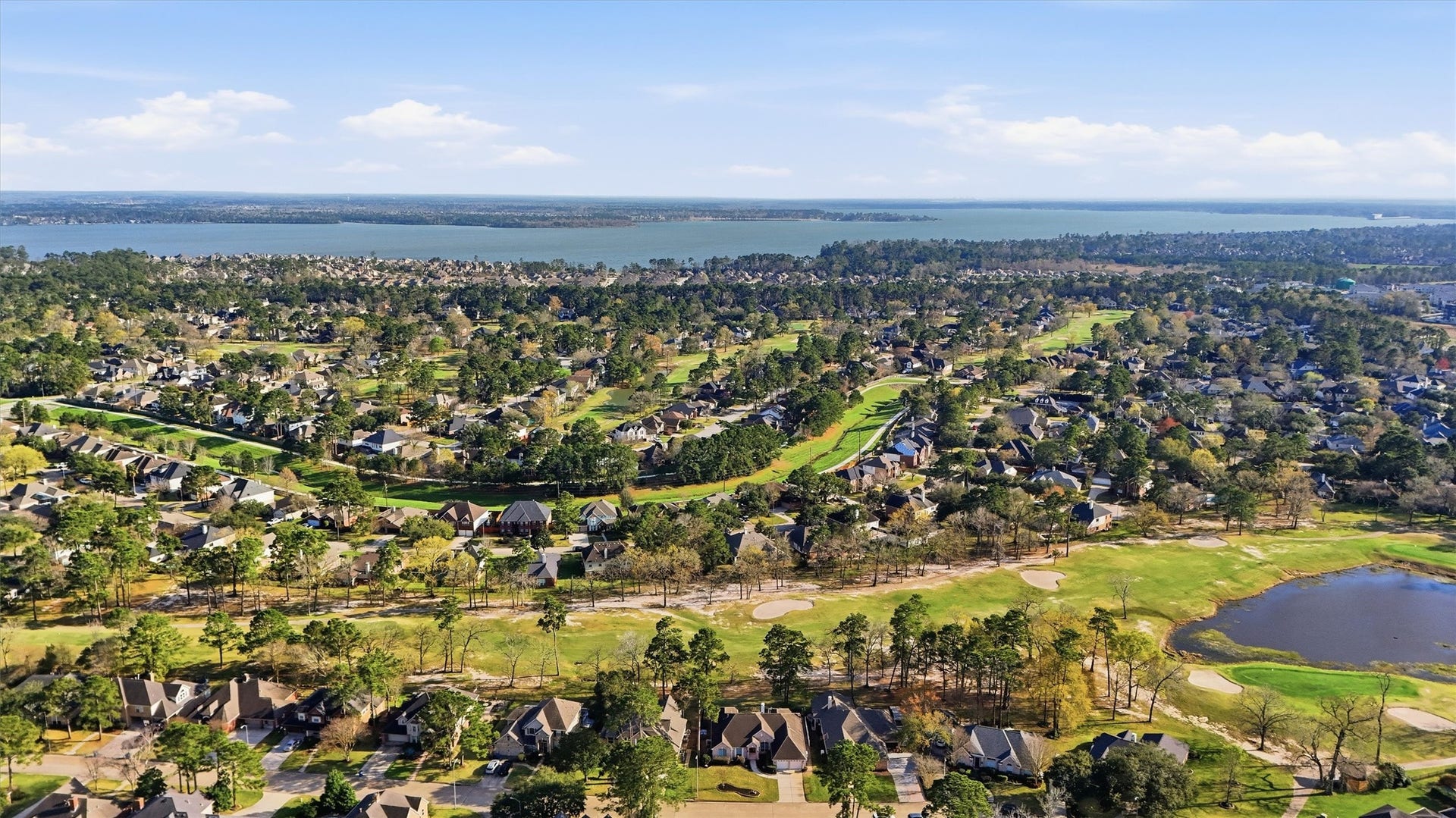 This aerial photo showcases a picturesque residential neighborhood nestled among lush greenery with a golf course and small bodies of water. In the distance, there's a scenic view of a large lake, offering serene and attractive surroundings for potential home buyers seeking a tranquil environment.