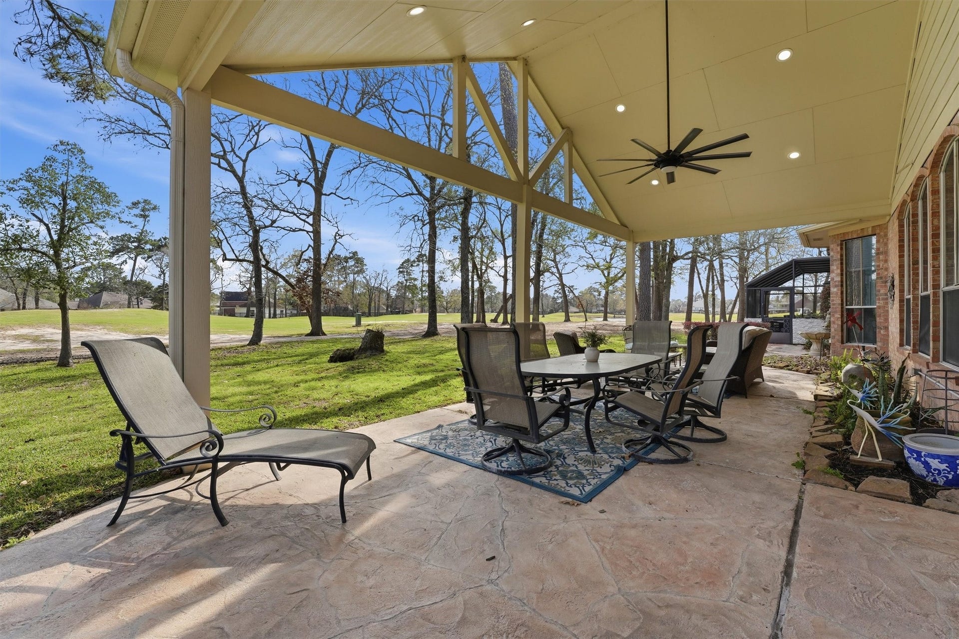 This photo showcases a spacious covered patio with a high ceiling, ceiling fan, and ample seating. It overlooks a lush, open yard with trees, perfect for outdoor dining and relaxation.  This house is located on the golf course, hole 3.